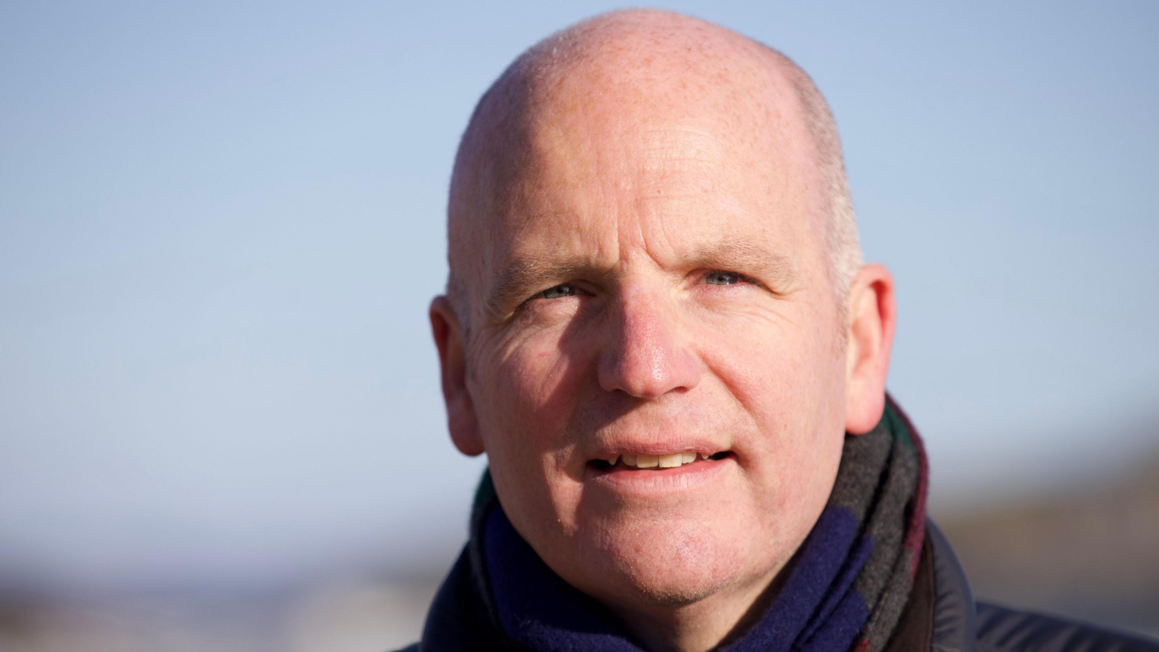 Glenn Campbell in close-up, looking into the camera. He can be seen wearing a dark blue and grey striped scarf.