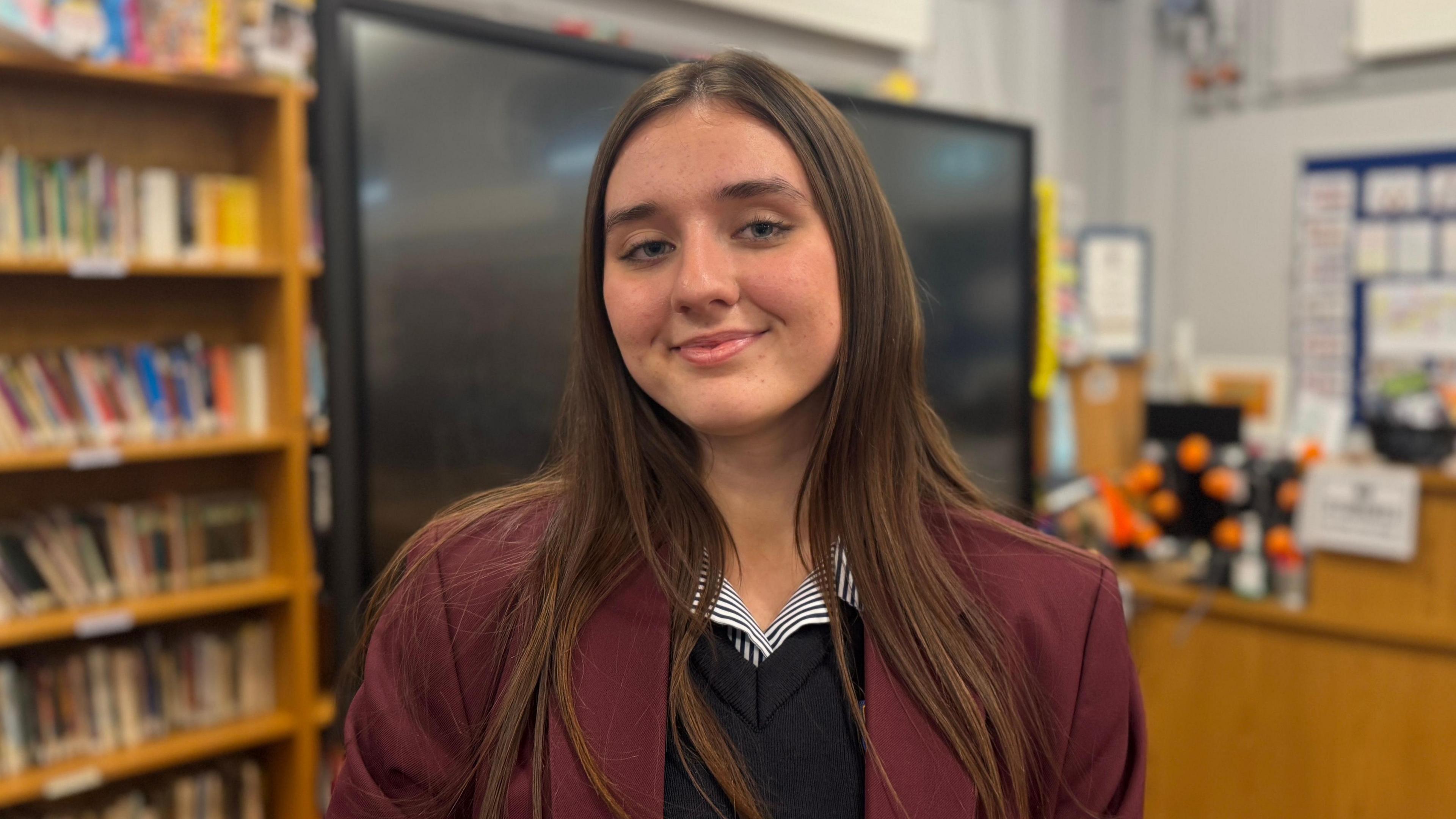 Rachel has long brown hair and is smiling at the camera. She is standing in the school library in front of a large screen. She is wearing a maroon school blazer, a striped shirt and a navy school jumper.