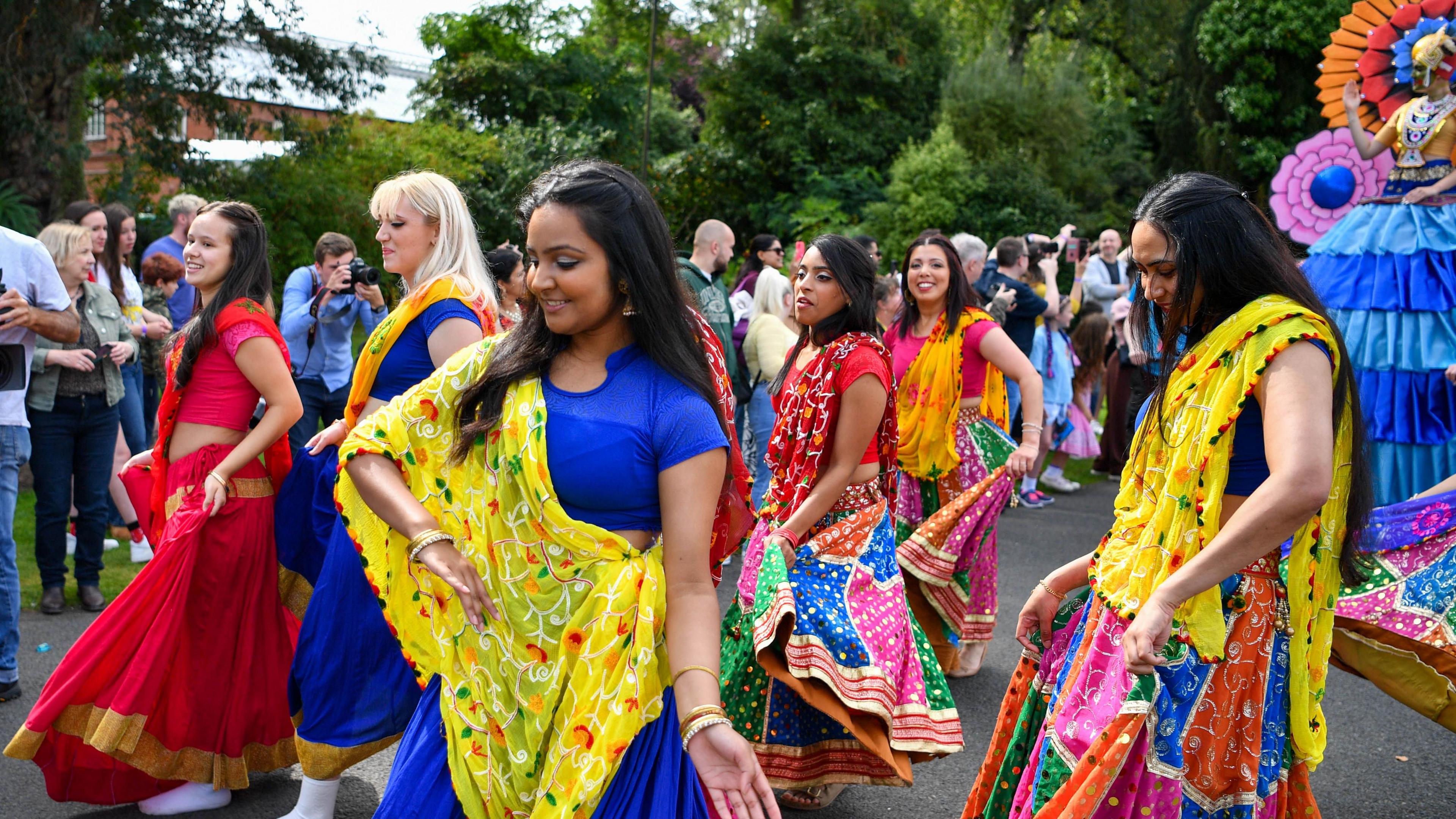 Women wearing bright colours are dancing in public gardens. Trees are in the background. Crowds are watching. 