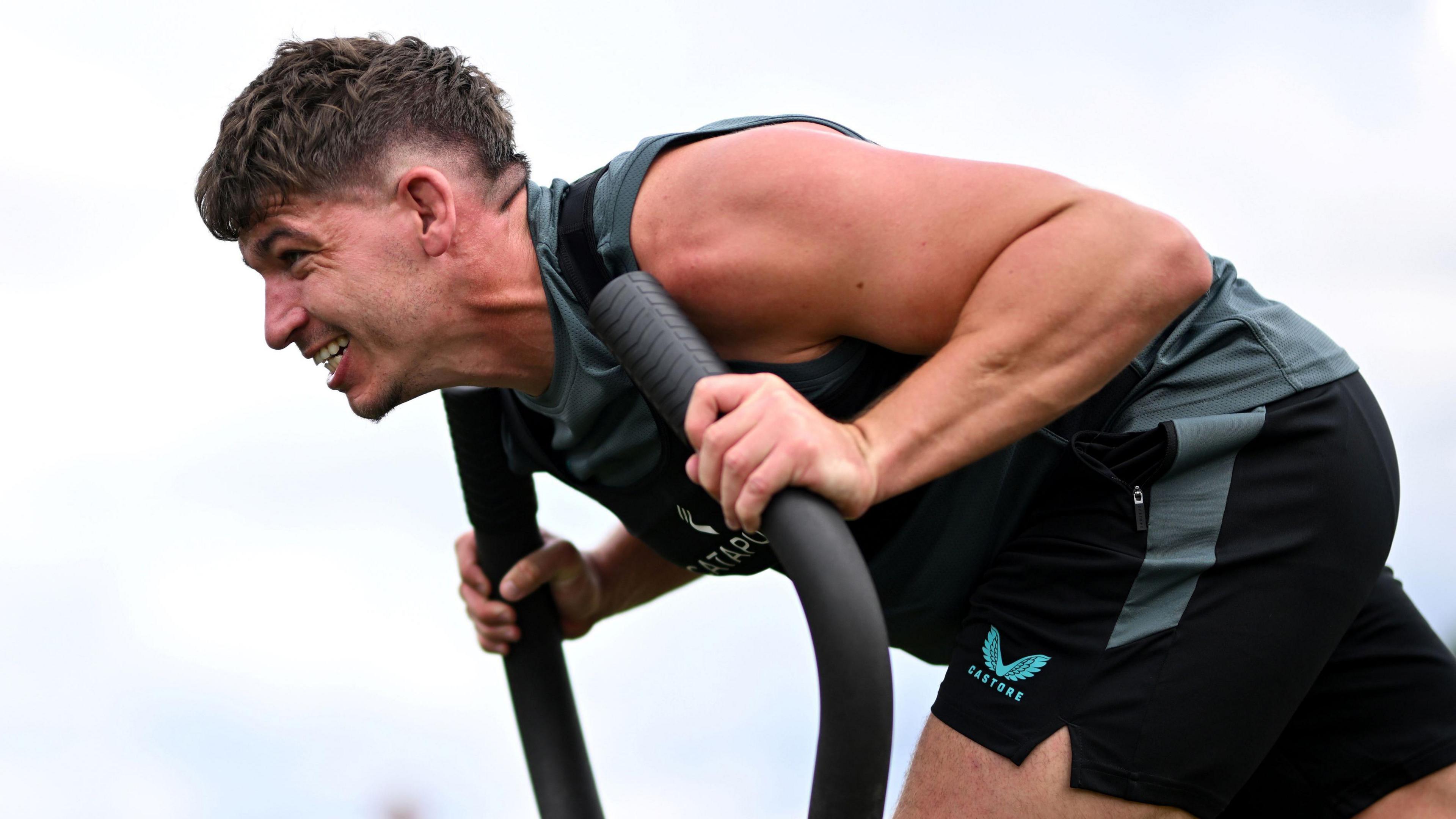 Rugby player Dan Frost bends forward as he pushes against a weighted sled during training. He is wearing black shorts, a black and grey vest, and looks like he is using a lot of effort. 