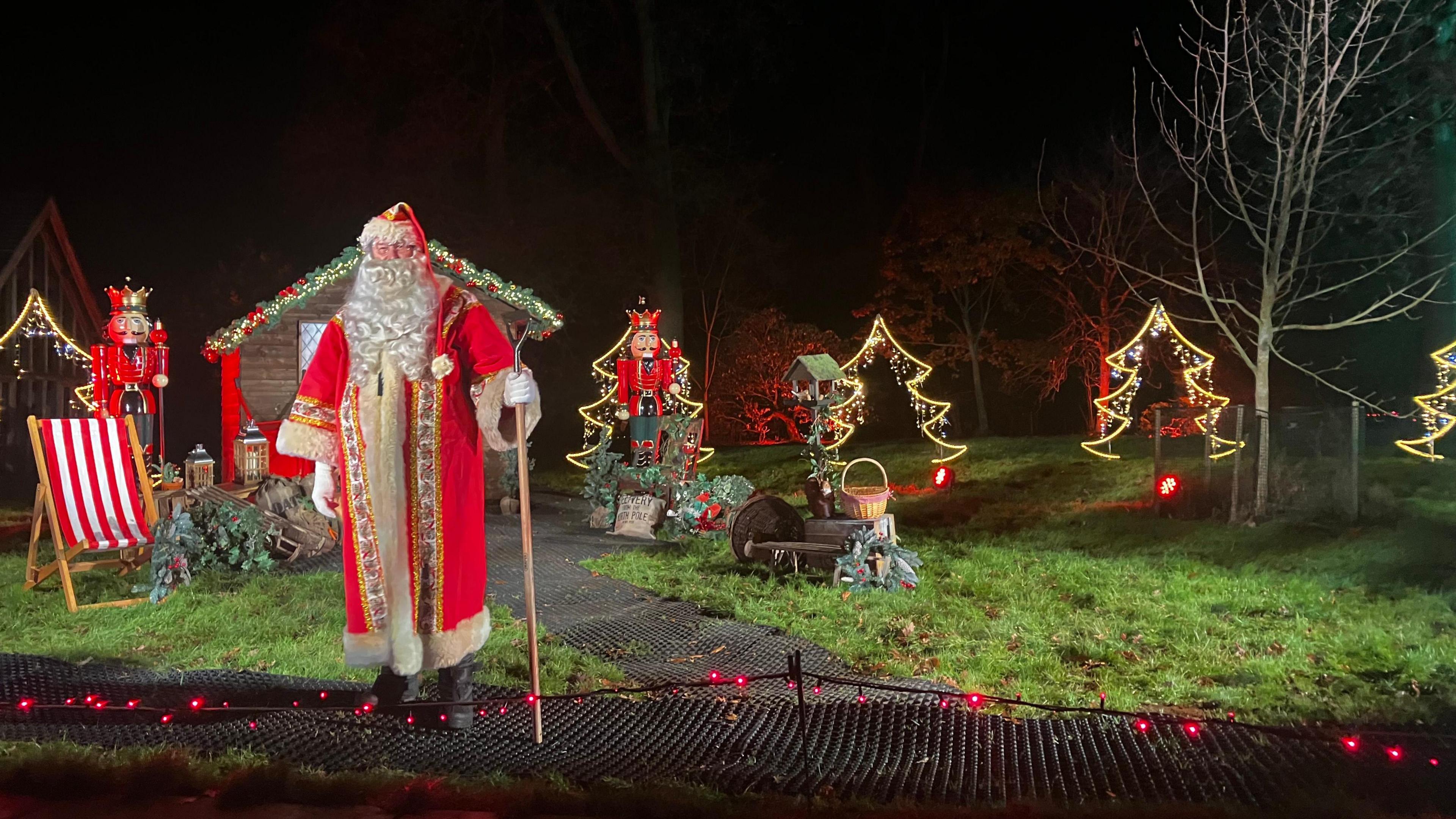 A Christmas scene at Westonbirt, comprising of a lifesize Father Christmas figure, two nutcracker soldiers, red and white stripy deckchairs, a festive shed, and lit up Christmas-tree shaped lights.