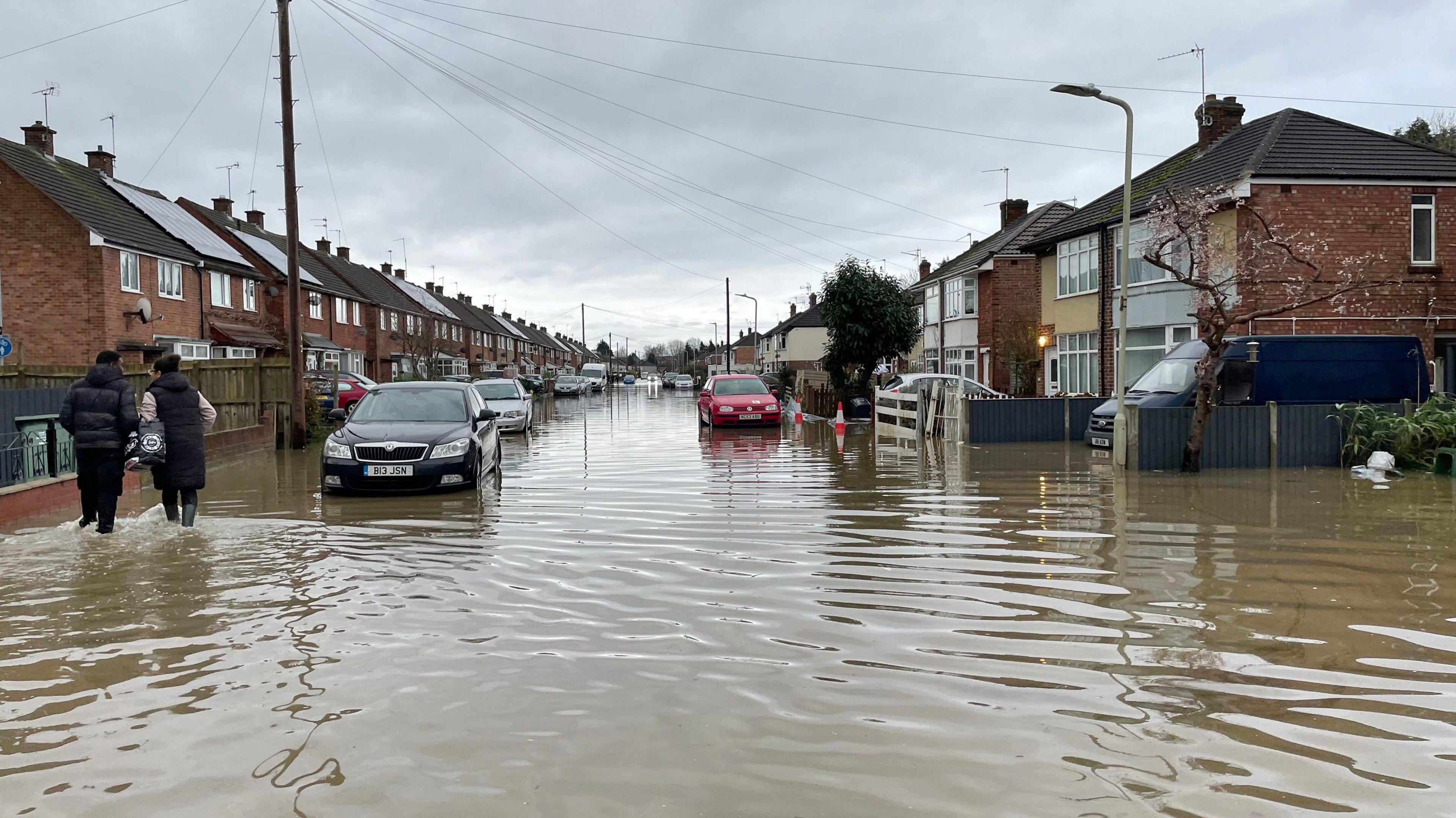 A flooded residential street with two people wading along the road in water up to the bumpers of cars