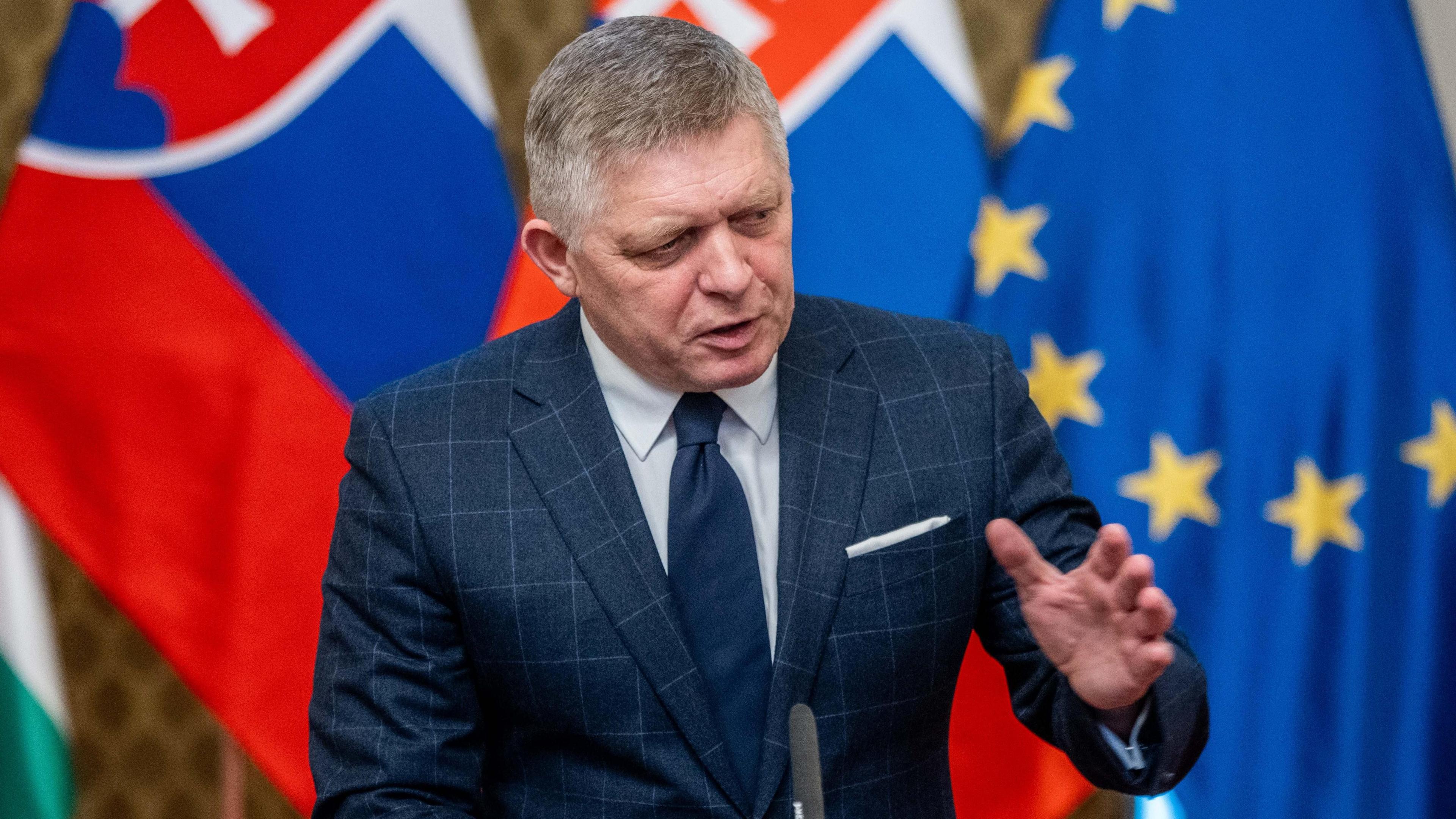 Robert Fico, wearing a blue checked suit and blue tie, gestures with his left hand as he holds a press conference on 21 January. Slovak and EU flags stand in the background