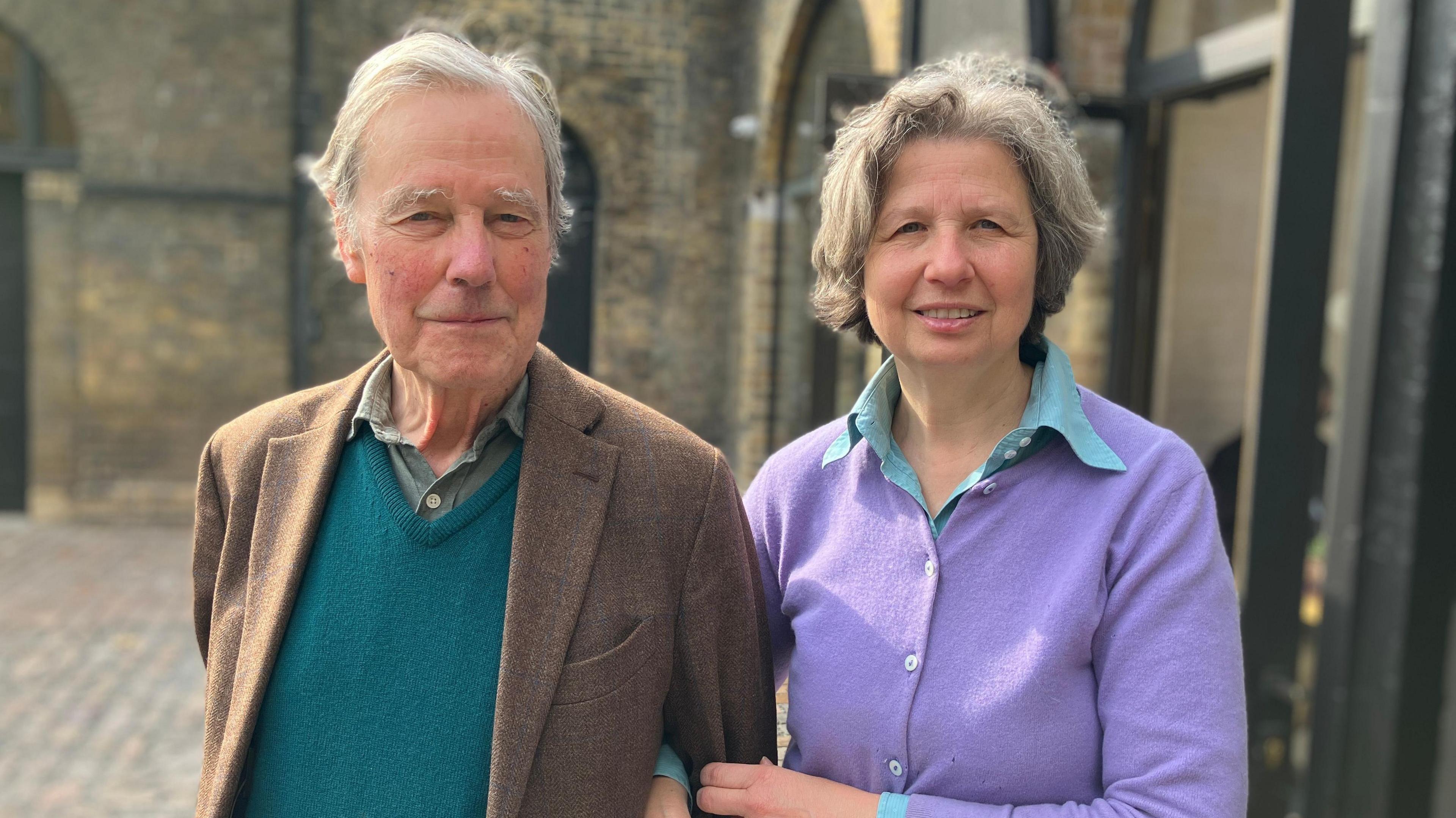 A woman with short grey hair on the right wearing a button-up lilac cardigan and a blue shirt underneath and linking her arm with a man wearing a brown blazer, teal v jumper and a green shirt. He has short white hair.