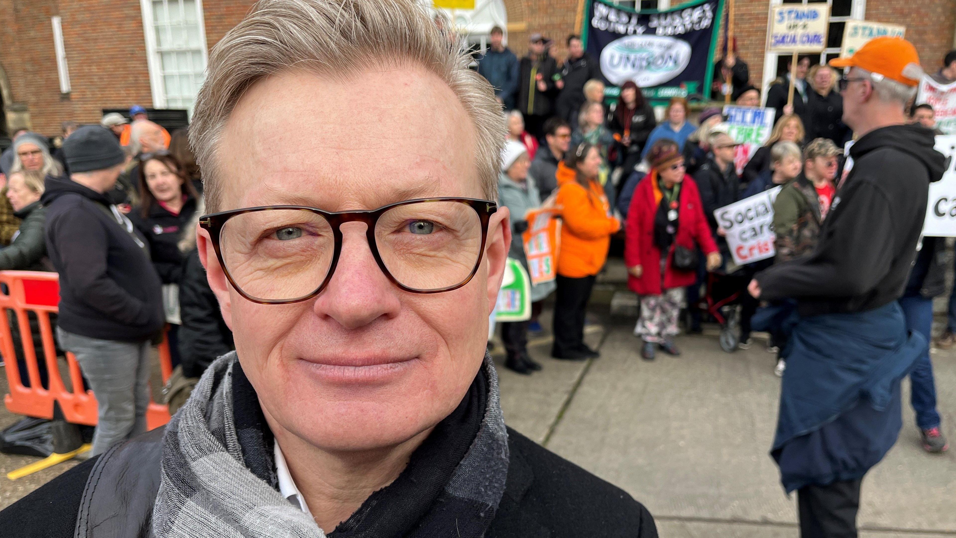 Neil Blanchard has short blond hair and wears glasses. He stand with the demonstration behind him wearing a black coat and a patterned grey scarf. 