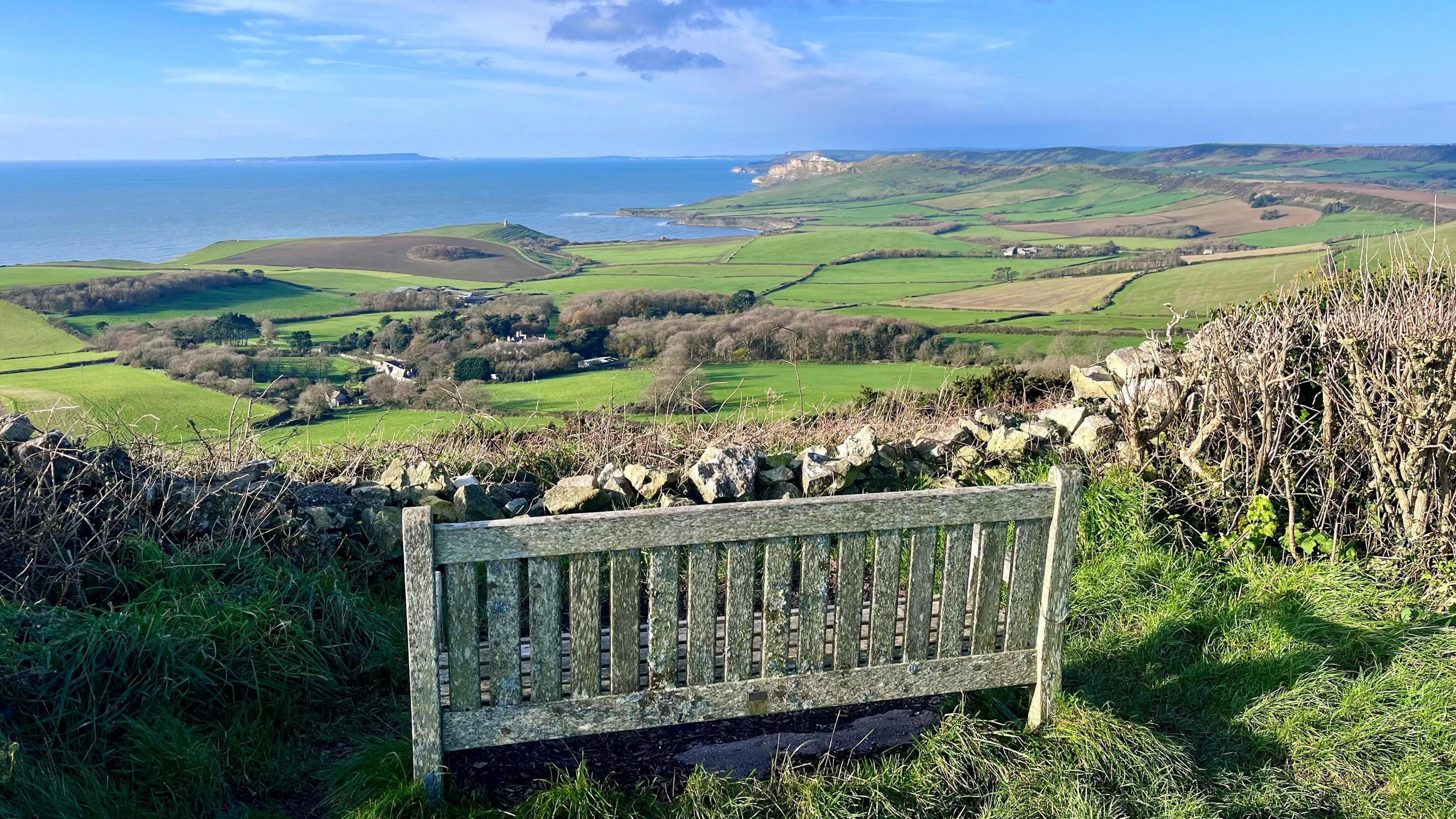 A wooden bench is in the foreground of the picture. Spread out before it is a patchwork of green fields until they reach the blue sea on the horizon. 