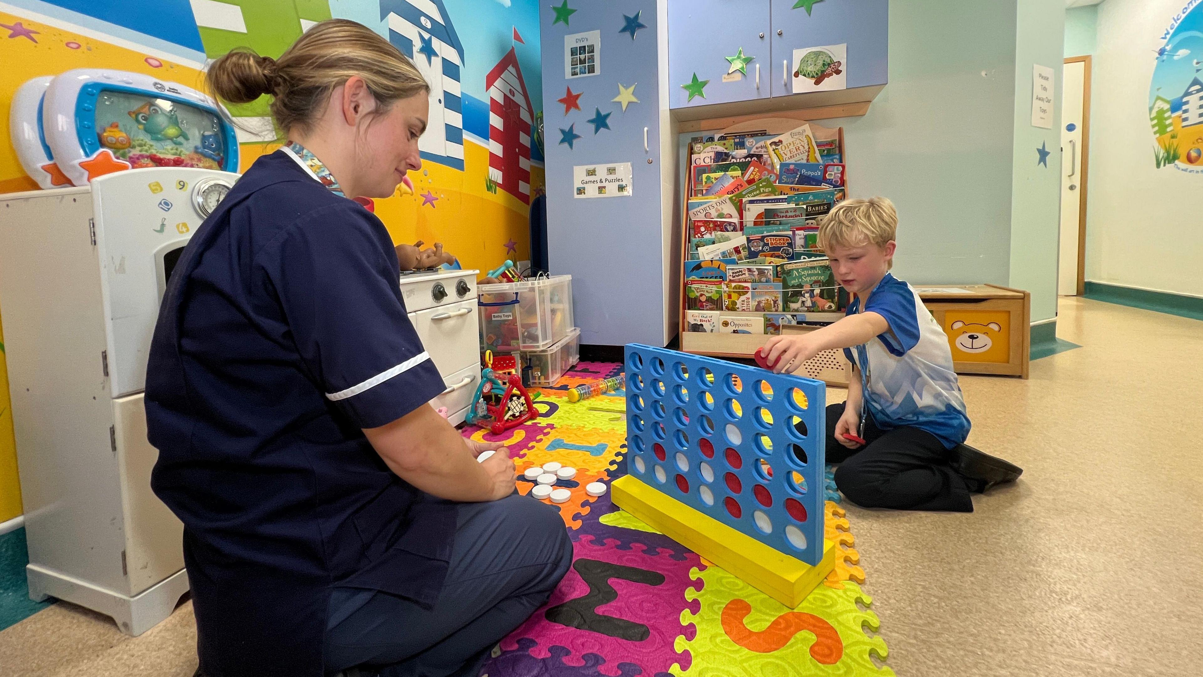 A boy playing Connect Four with a nurse in the toy room at the children's hospital ward. He is about to put a red token into the game as the nurse watches him. Alfie is wearing his race top from the Marathon Mile and his medal. The nurse is in her uniform and is kneeling in front of the game.