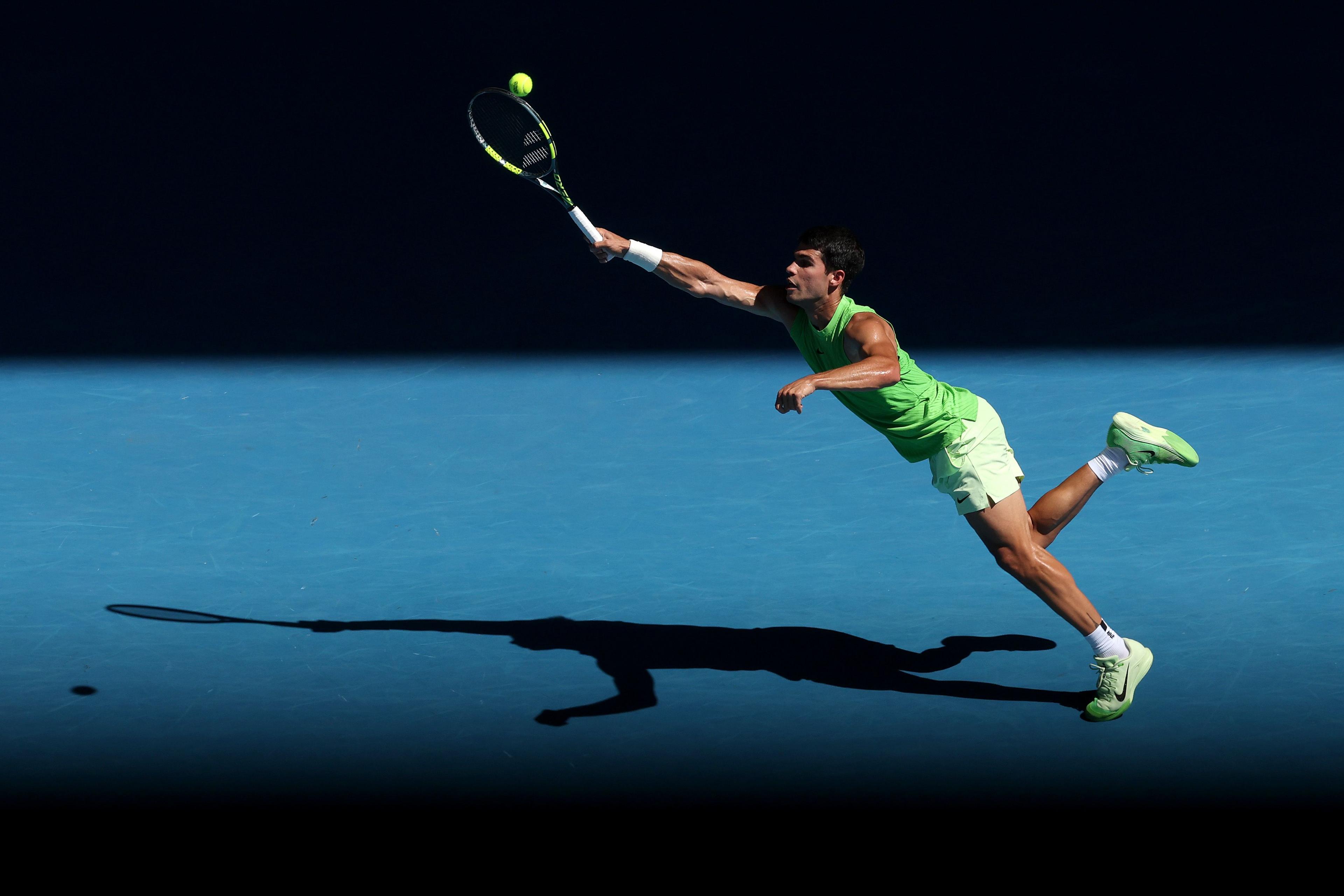 Spanish tennis men's world number one Carlos Alcaraz leaps forward on a blue hard court, fully extending their arm and racket to reach a high ball under strong sunlight, casting a long shadow behind them. 