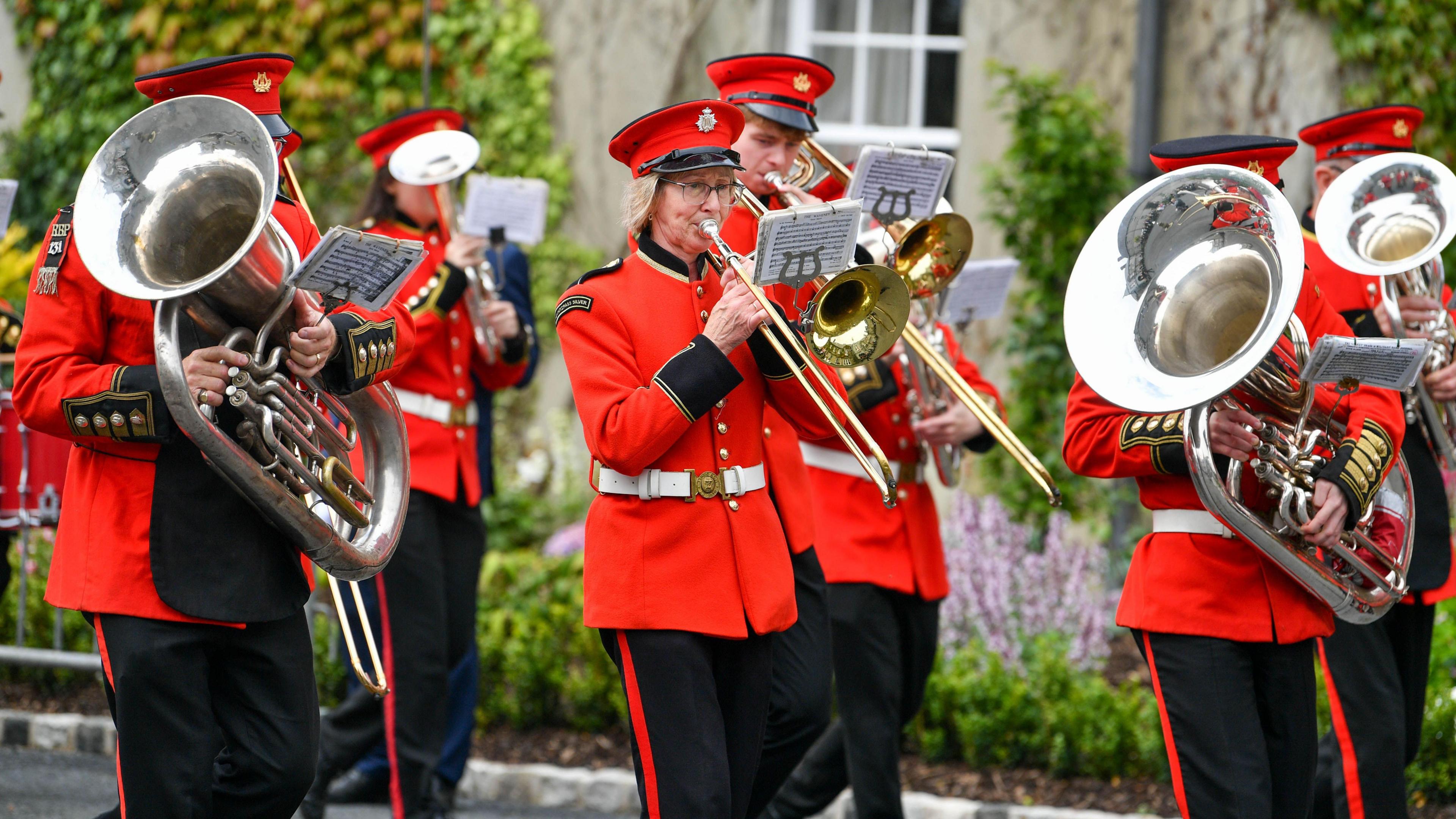 A band playing brass instruments wear a red jacket, white belt and black trousters. All members are wearing a red, flat top hat as they play outside a grey house.