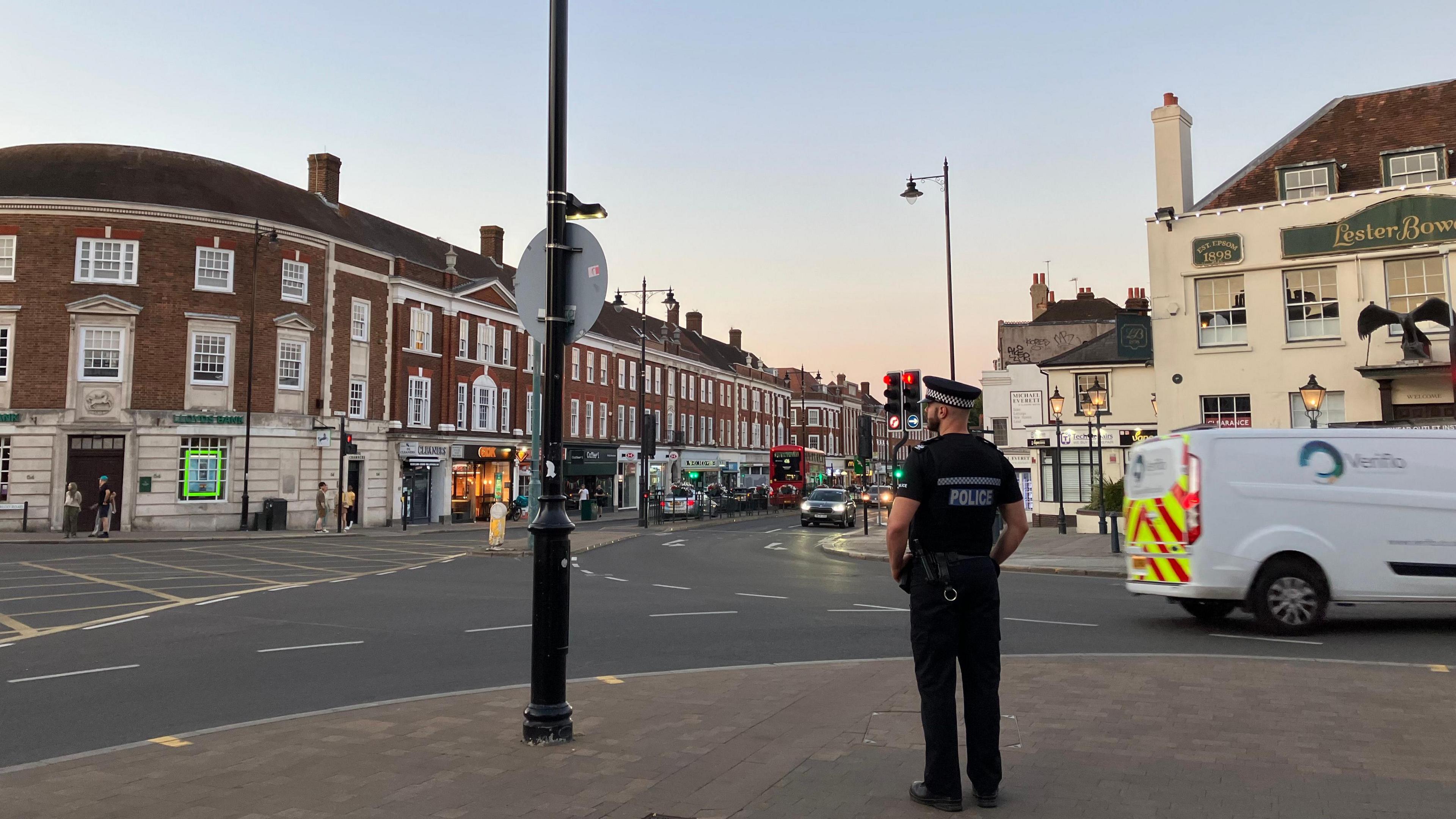 A police officer stands with his back to the camera on a street corner in Epsom. The road visible in front of him has three storey buildings along it with shops at ground floor level, and it is early evening.
