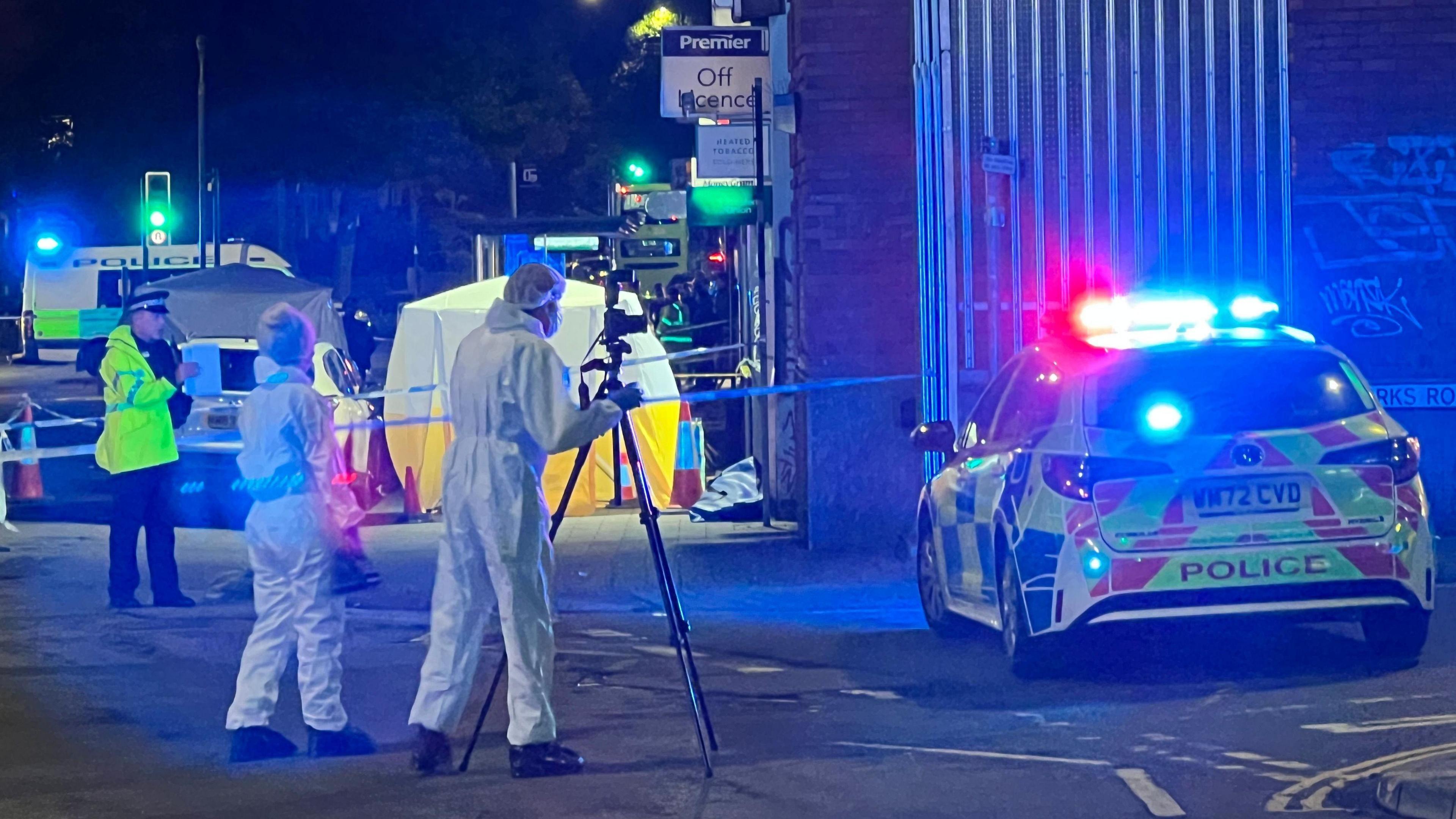 A forensic team dressed in white suits taking pictures using a tripod at the crime scene on Stapleton Road. There are police cars parked next to them with their lights on, and a white and yellow forensic tent is positioned outside the entrance to an off-license.