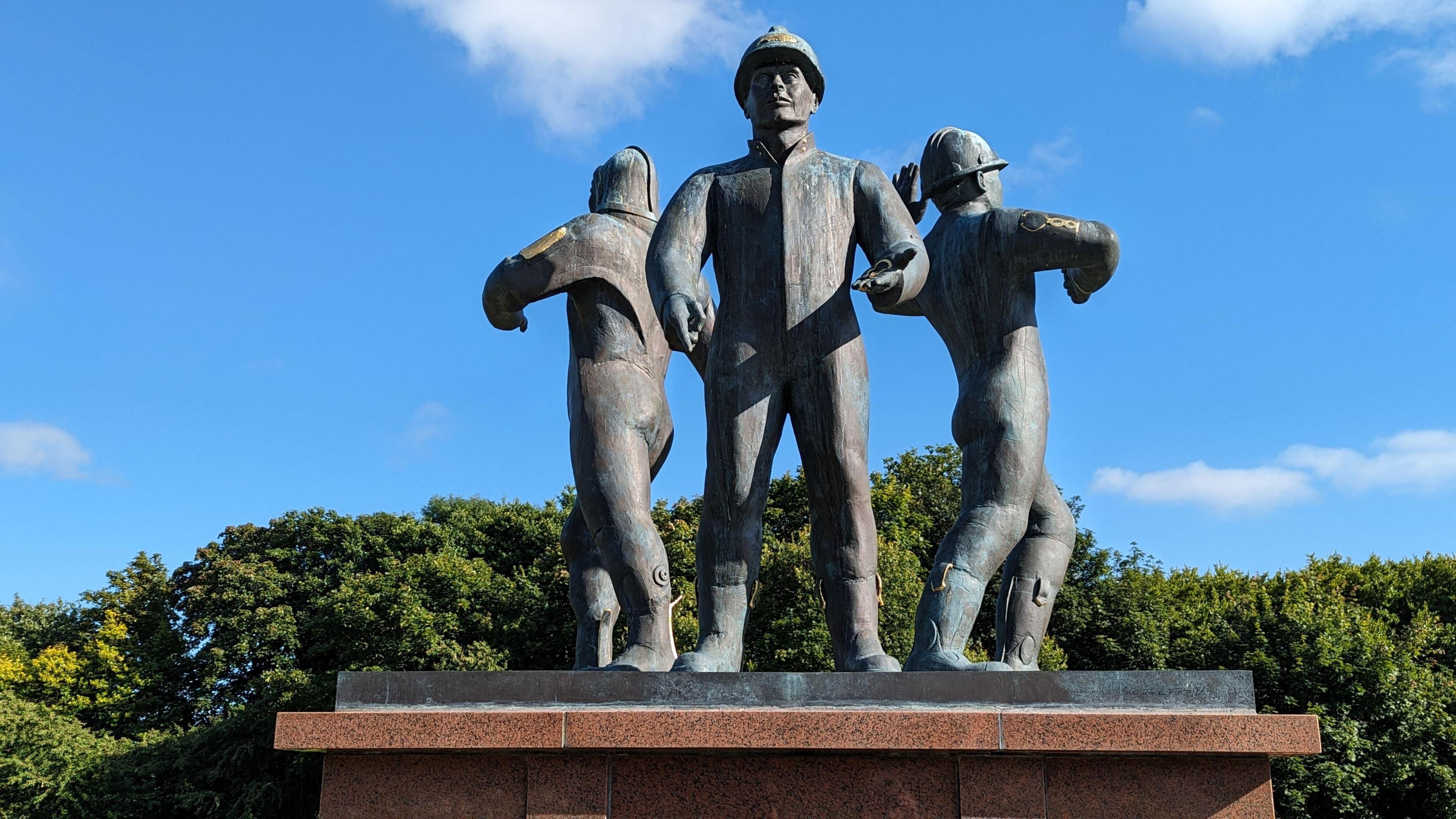 A blue sky and a horizon of green trees in full bloom. In front, a bronze statue of three oil workers in boiler suits and helmets, back-to-back and facing three directions. They are on a red granite plinth.