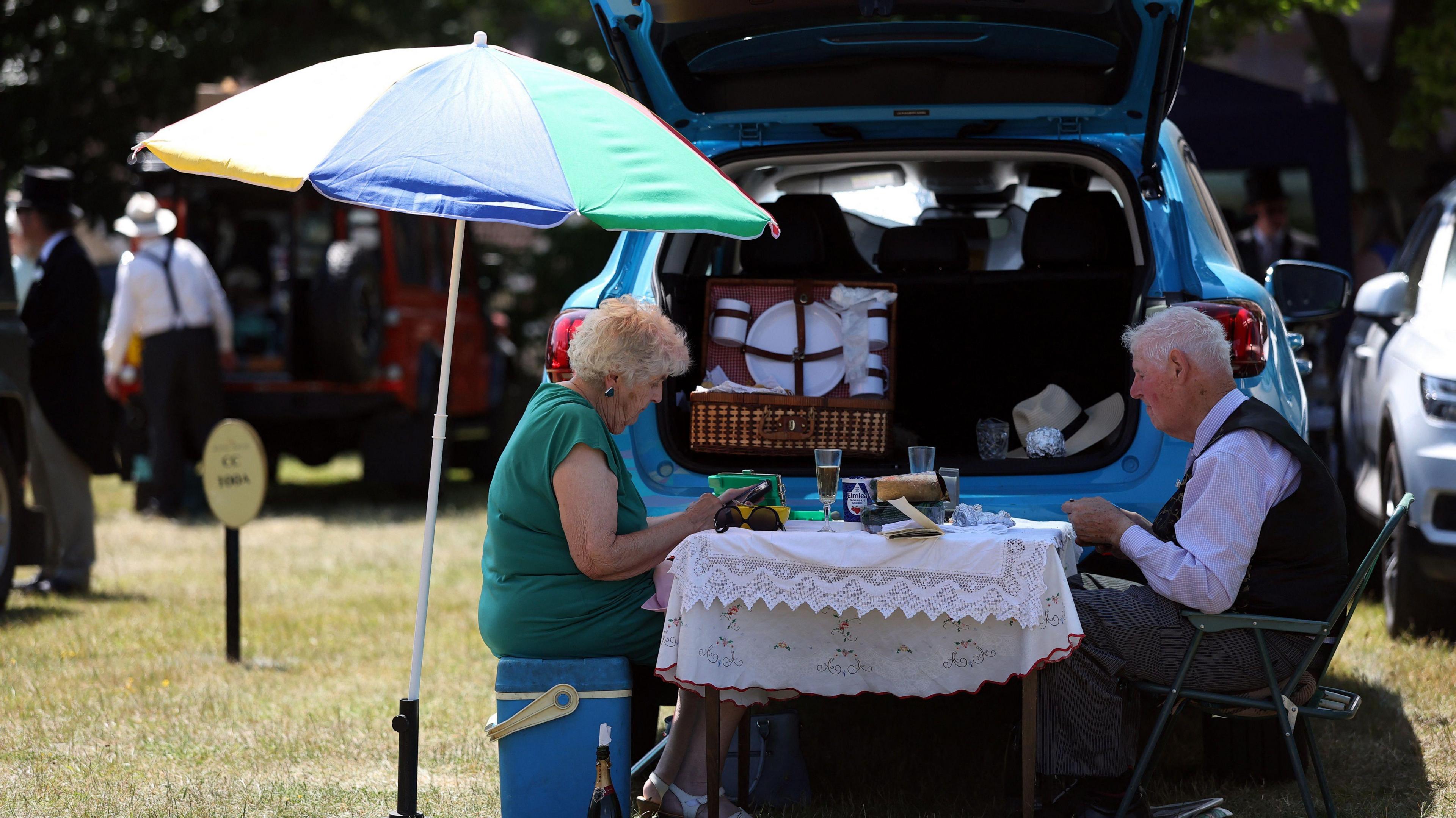 A man and a woman are sat at a table at the back of their blue car eating lunch. The table has a white tablecloth laid across it and there is a picnic set in the car's boot.