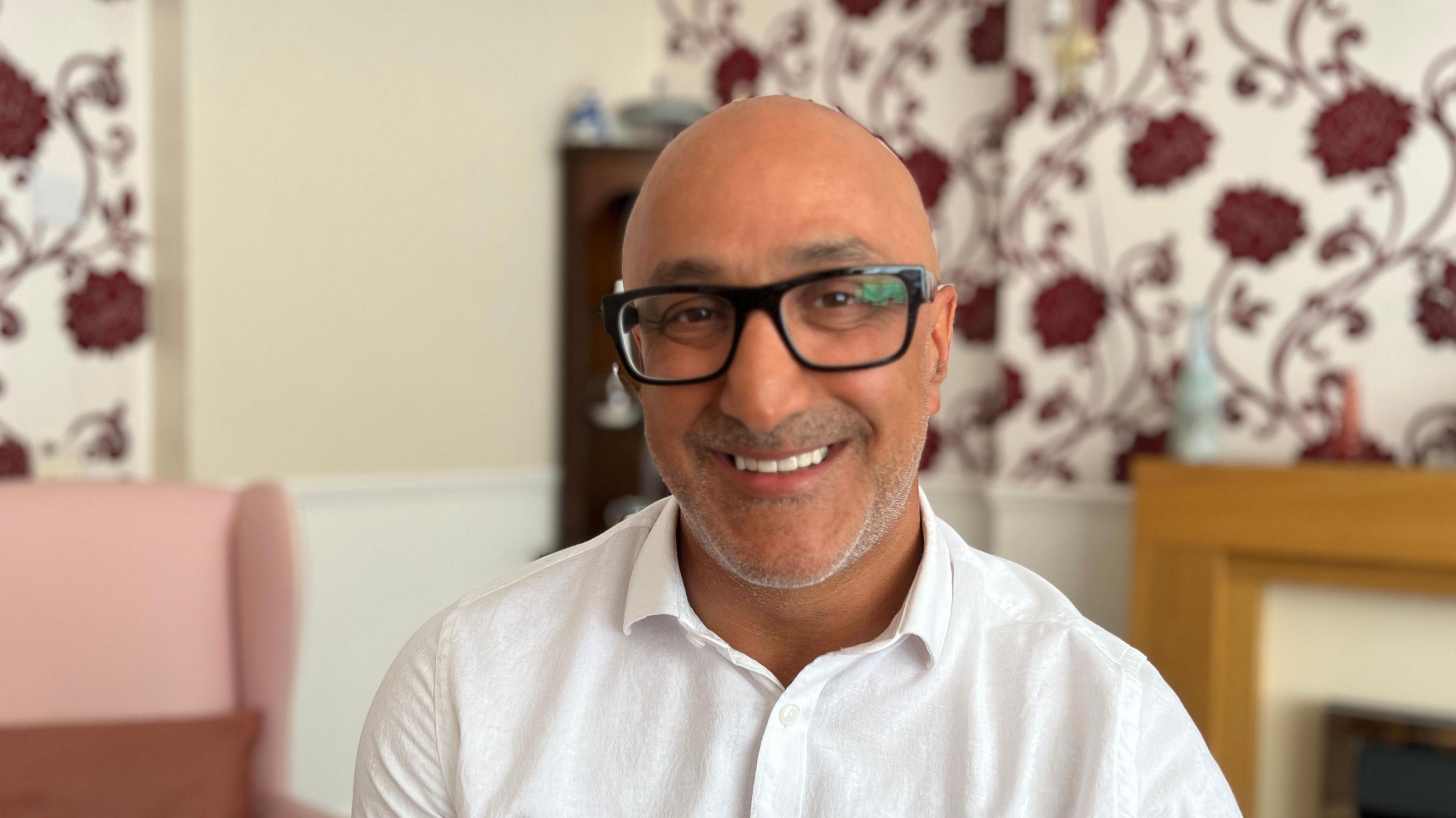 A middle-aged man with thick black-rimmed glasses is smiling at the camera, wearing a white shirt and sitting in a living room.