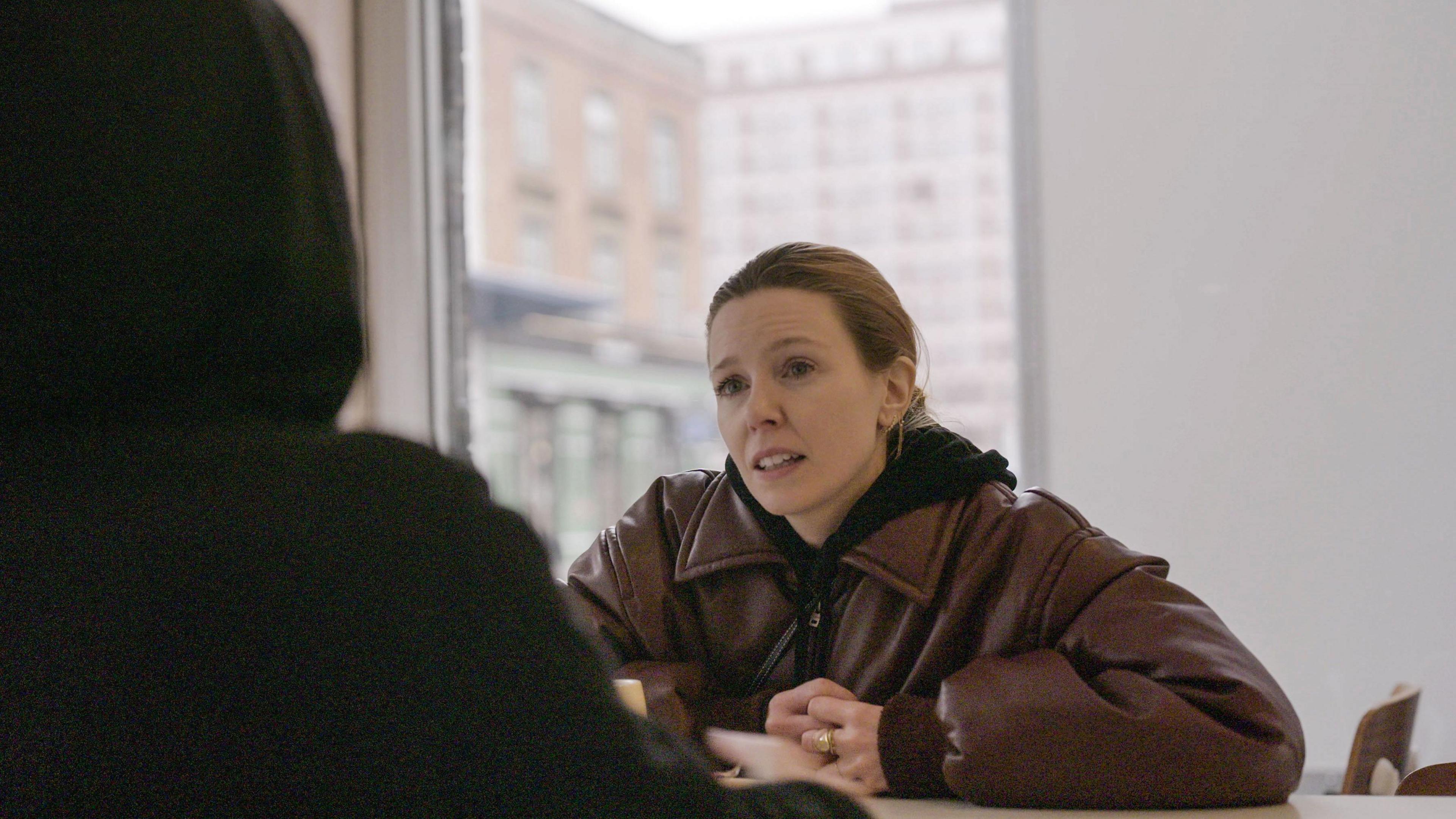 Stacey Dooley sits opposite a person in a black hoodie. A woman with ginger hair and a brown leather jacket sits opposite a person in a black hoodie. 