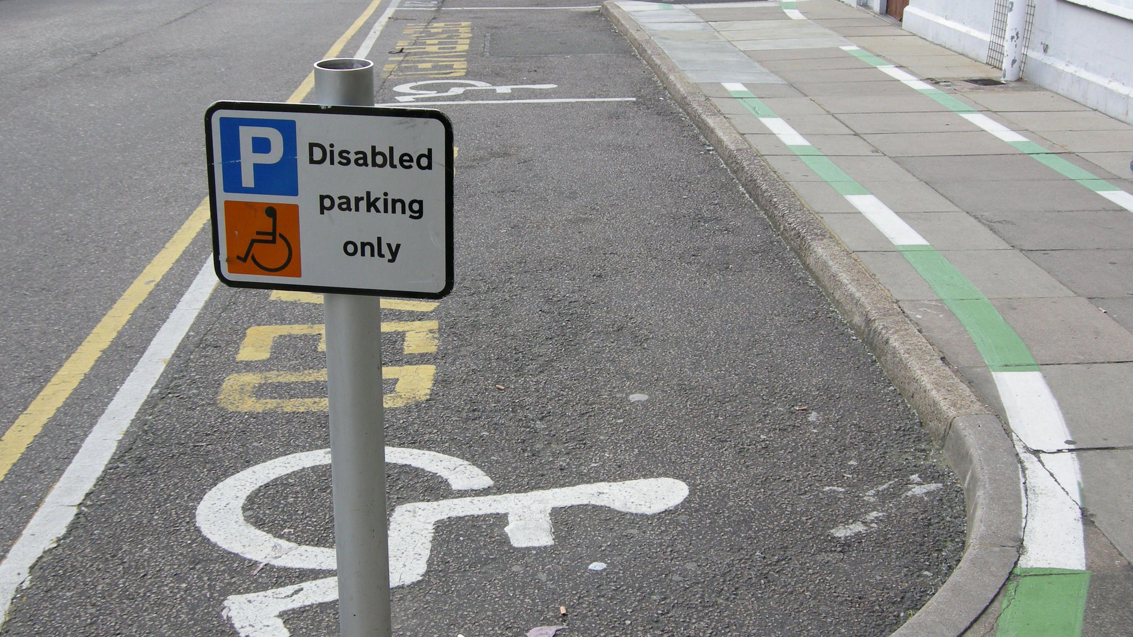 A disabled parking bay with markings painted on the road and a sign on a short metal pole. There is a wheelchair drawn on the road and on the sign.