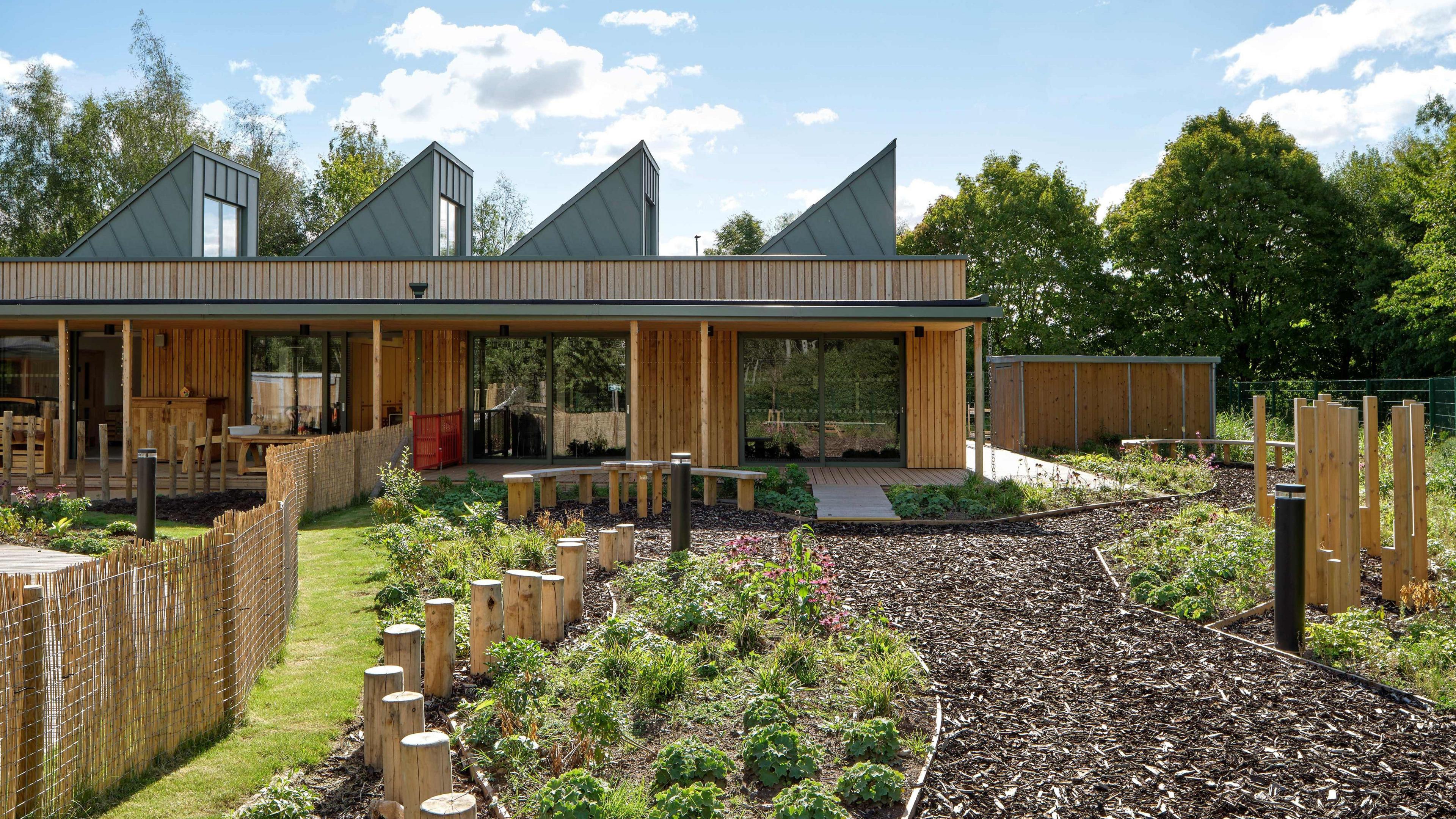 A garden with a bark path and wooden planters, with a wooden triangular building in the background. There are also trees in the background and small plants in the garden.