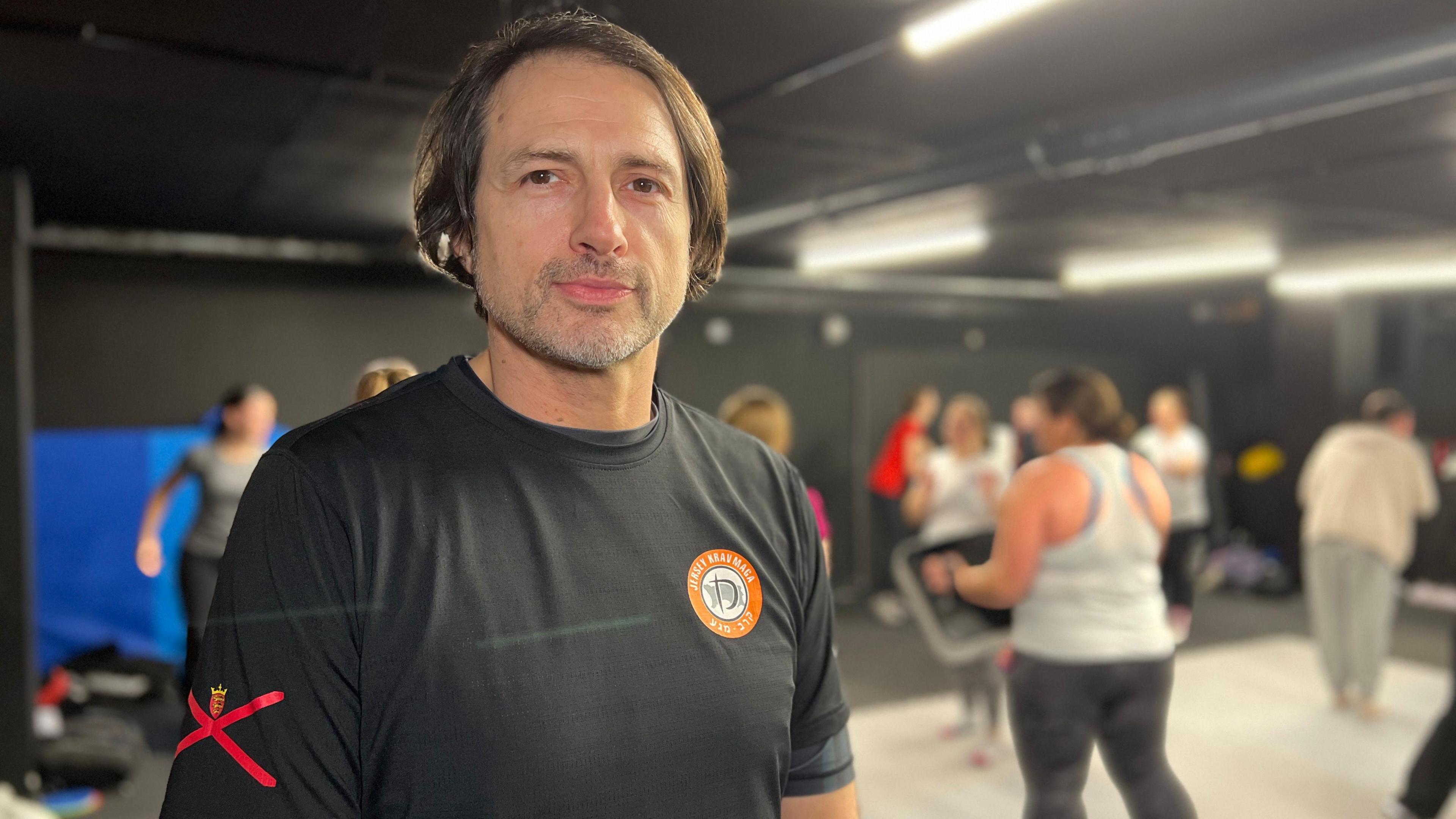 Richard looks at the camera while the girls train behind him holding pads. He is wearing a black exercise top with a black t-shirt which has the club's logo on his chest and the Jersey flag on his sleeve. Richard has floppy brown hair and brown eyes.