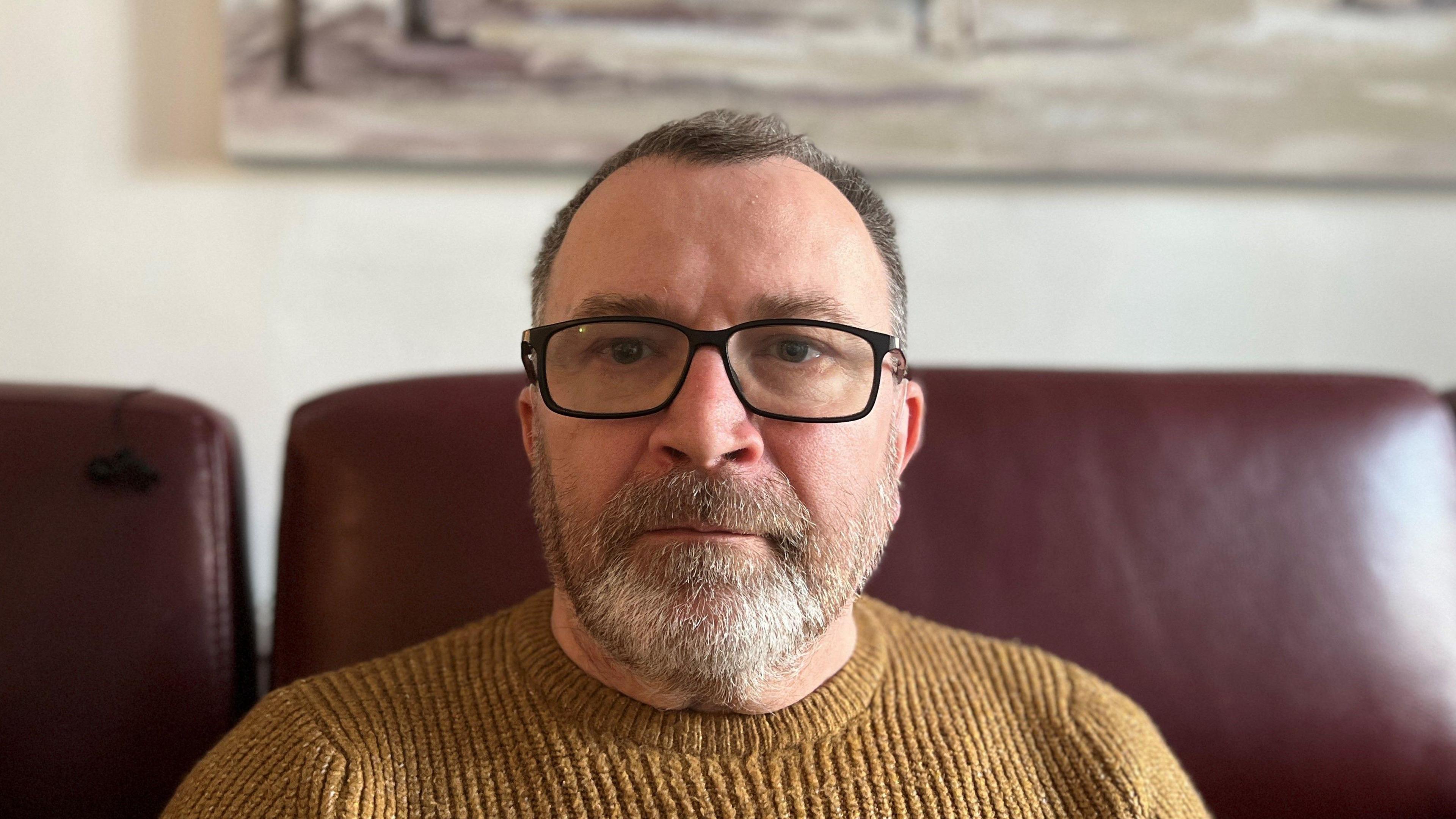 Head and shoulders shot of Christopher Price, sitting on a brown leather sofa wearing a light brown jumper