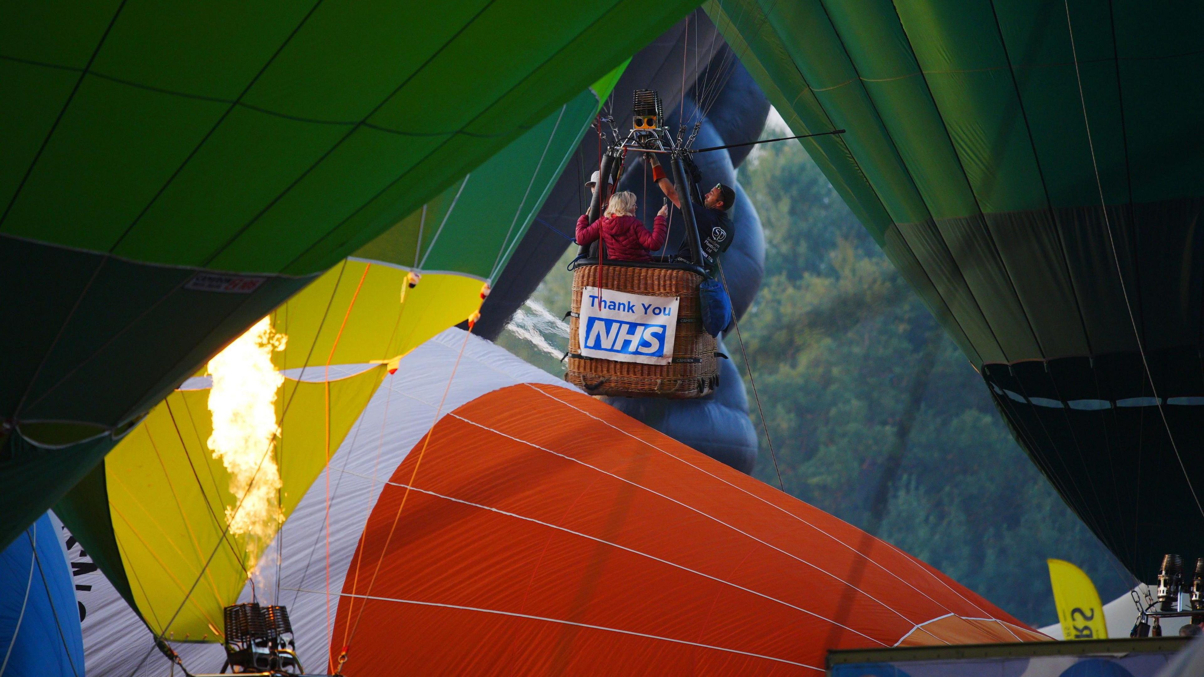 Passengers in a hot air balloon are beginning to take off, surrounded by other balloons. On the outside of the wicker basket is a thank you NHS sign.