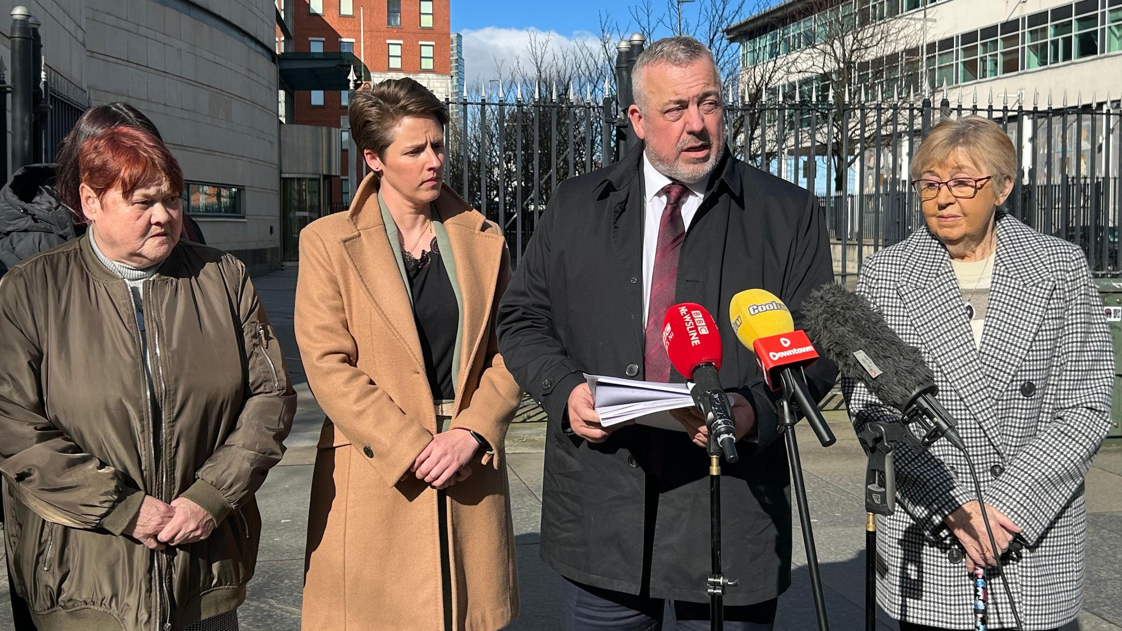 Four people standing outside Belfast Crown Court. A man wearing a coat, shirt and tie speaks at microphones while holding a notepad.  Standing on either side of him are three women