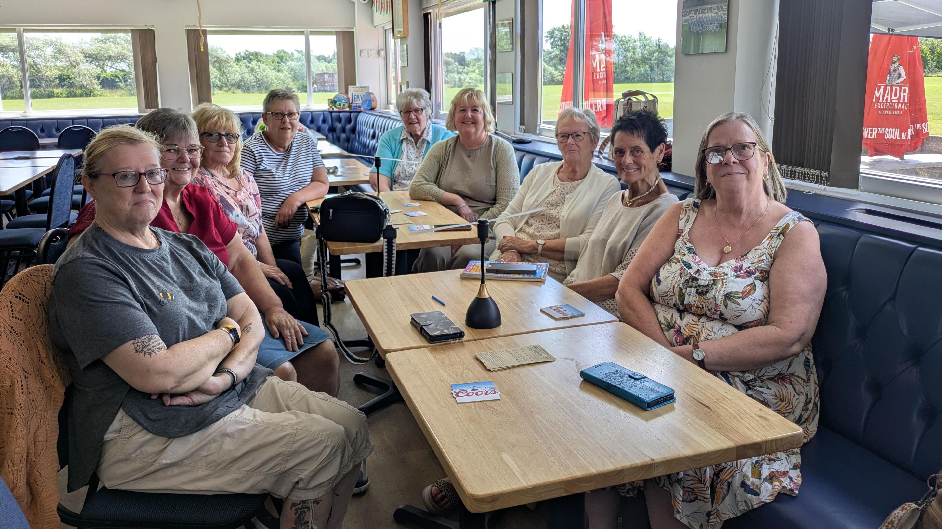 A group of women sitting around two brown tables having a coffee morning in a sports club 