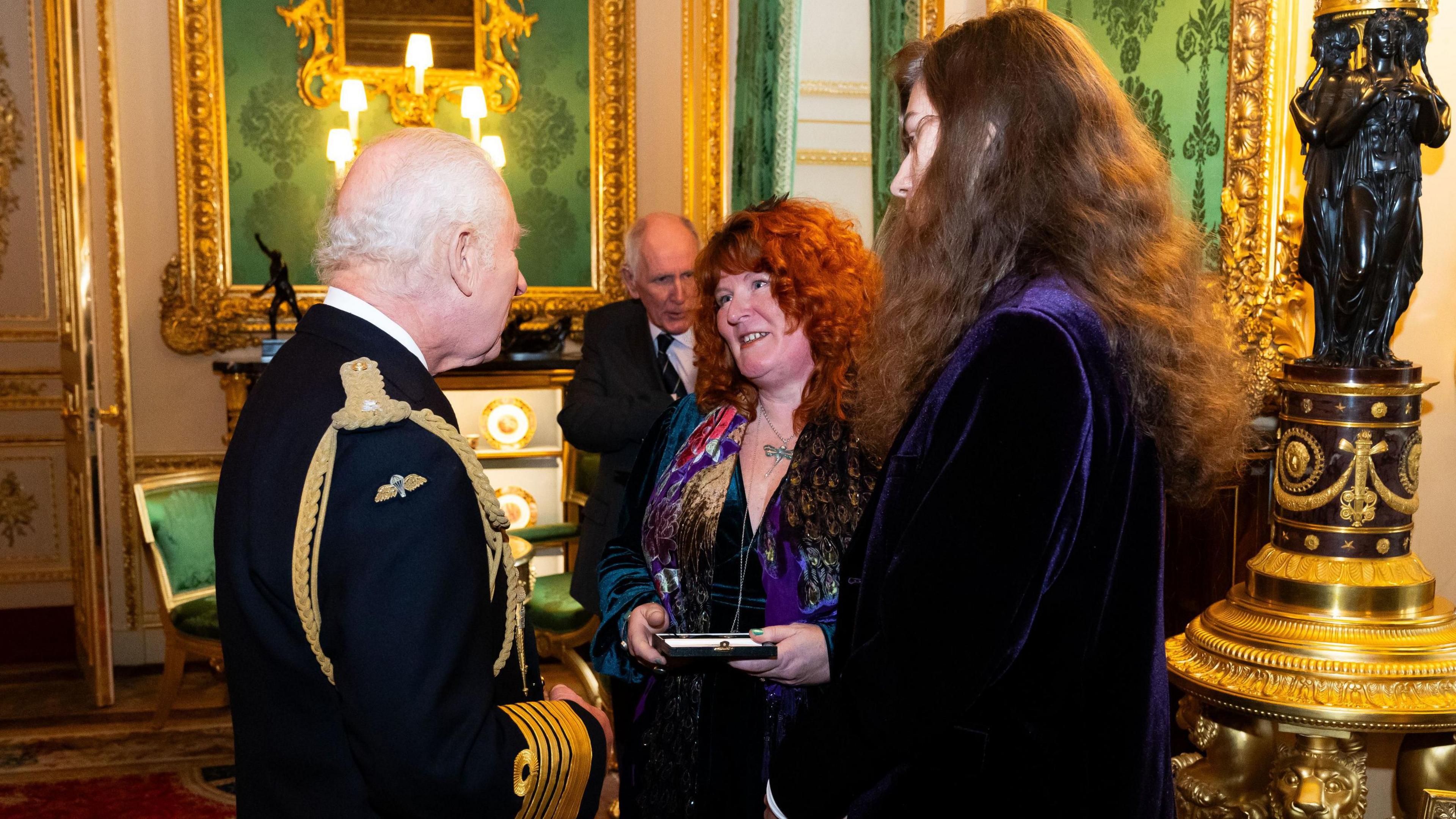 King Charles III presents an Elizabeth Emblem to Rebecca Lombard-Earl. They are in a gold and green grandly decorated room. Her son Rowan is also with her,