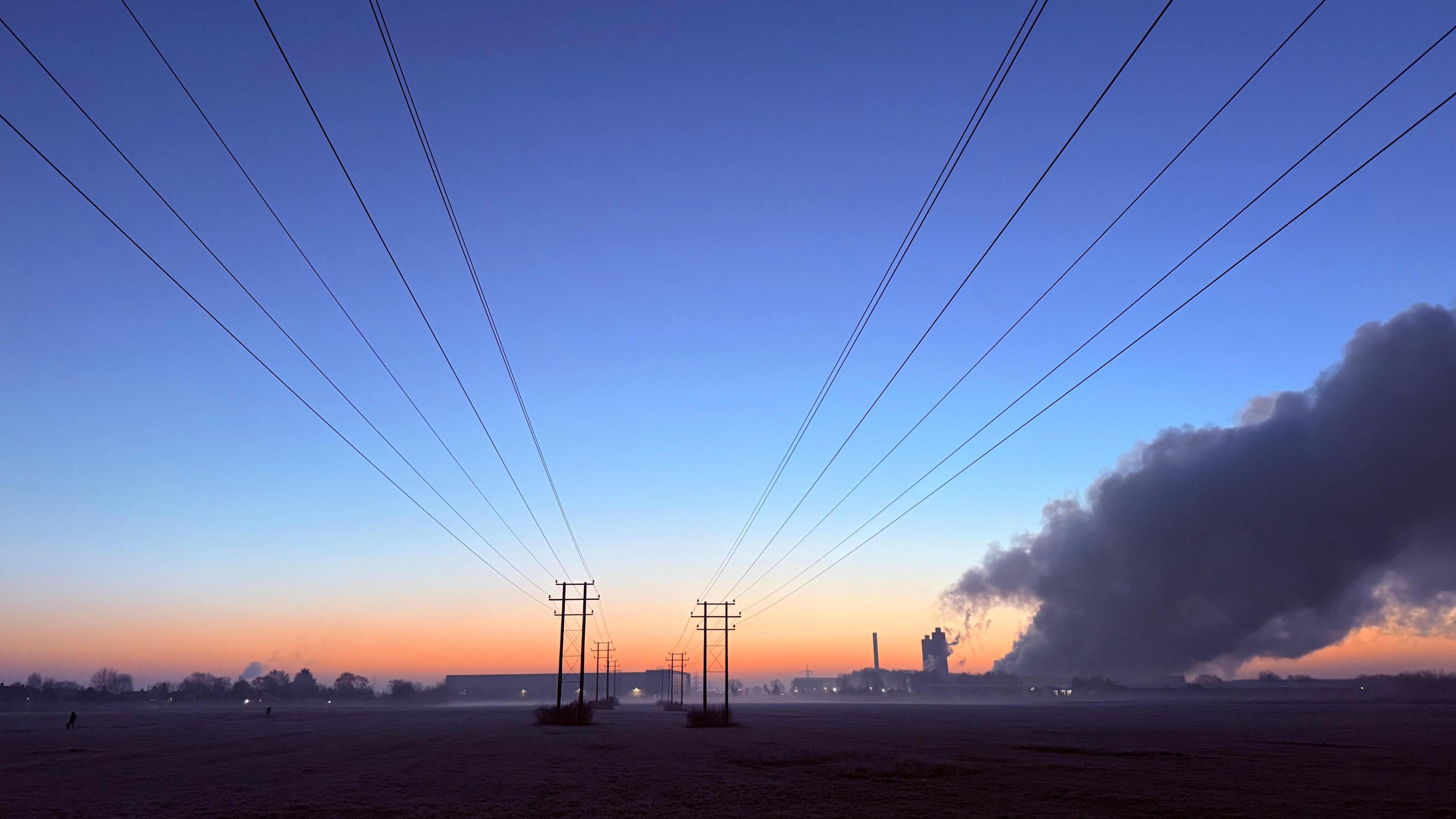 A vivid blue sky glows orange on the horizon at sunrise. Power lines run through the centre of the photo along a line of telegraph poles. In the distance you can see large plumes of smoke coming from a buildings large chimney. 