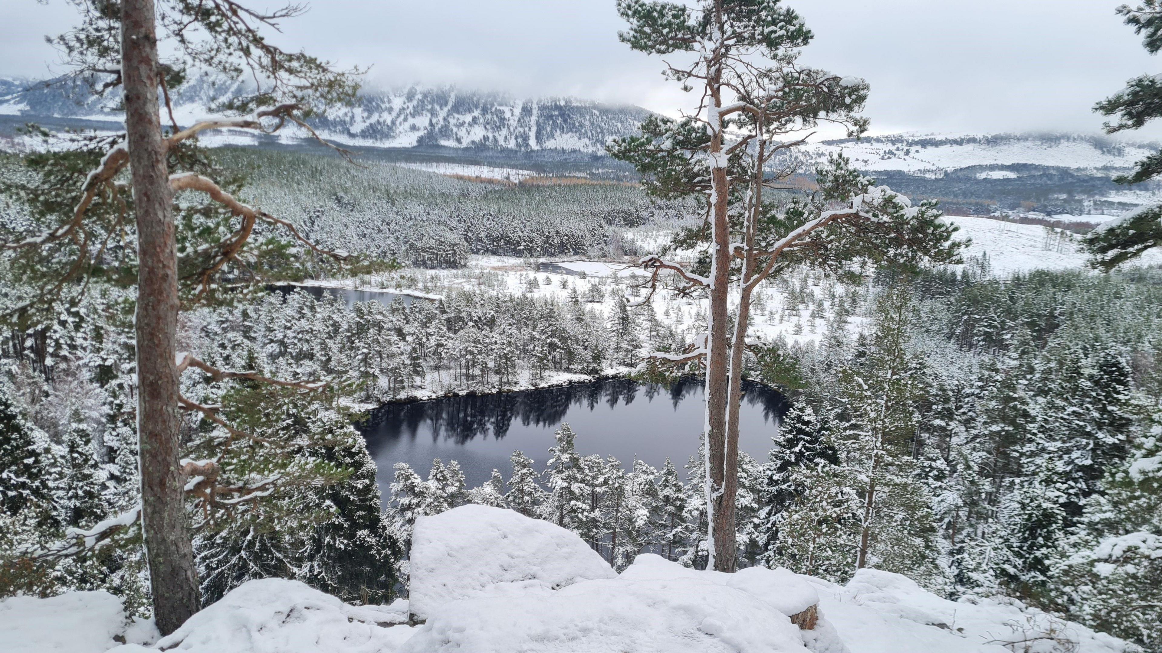 Snow-covered landscape with pine trees and a dark lake surrounded by snowy forest and distant mountains.