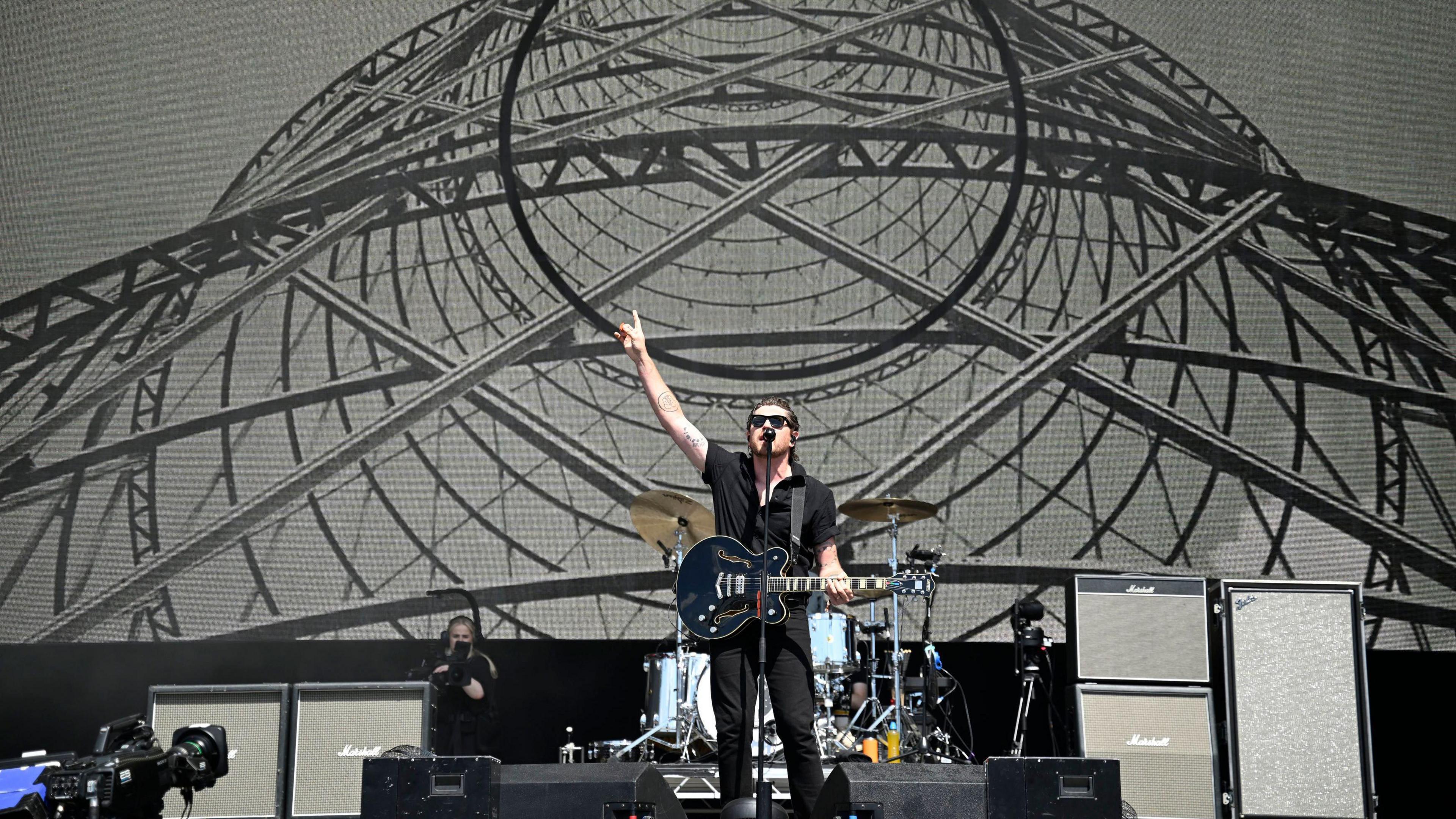 A wide shot of a large stage. The lead singer is all in black, playing what appears to be a black Gibson semi-hollow body guitar. He is punching the air and singing. Dark glasses shield his eyes from the sun. There is a photo backdrop of what appears to be TV transmission aerial.