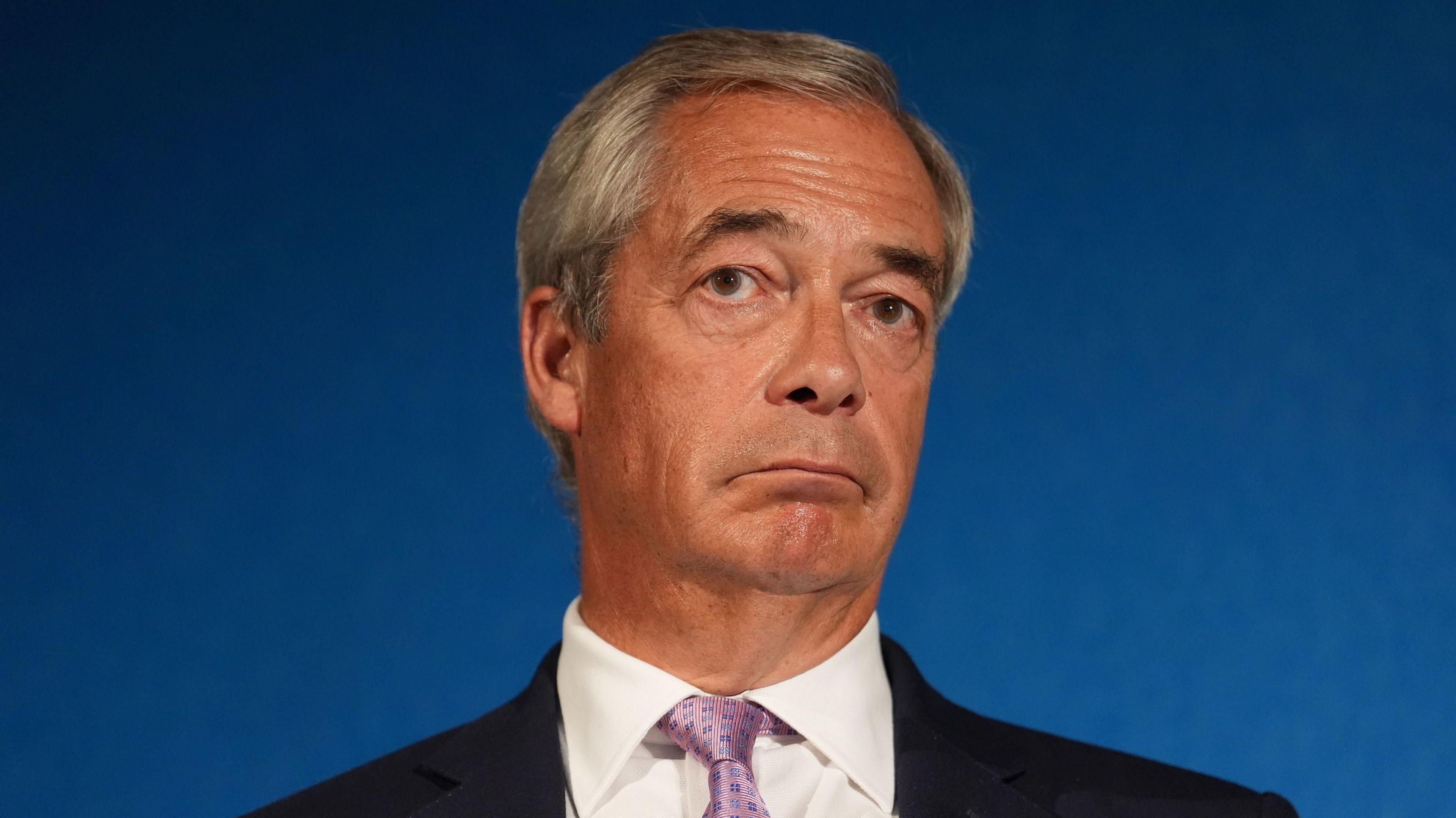 Reform UK party leader Nigel Farage looks stern at press conference, wearing a black suit jacket, white shirt and a pink tie.