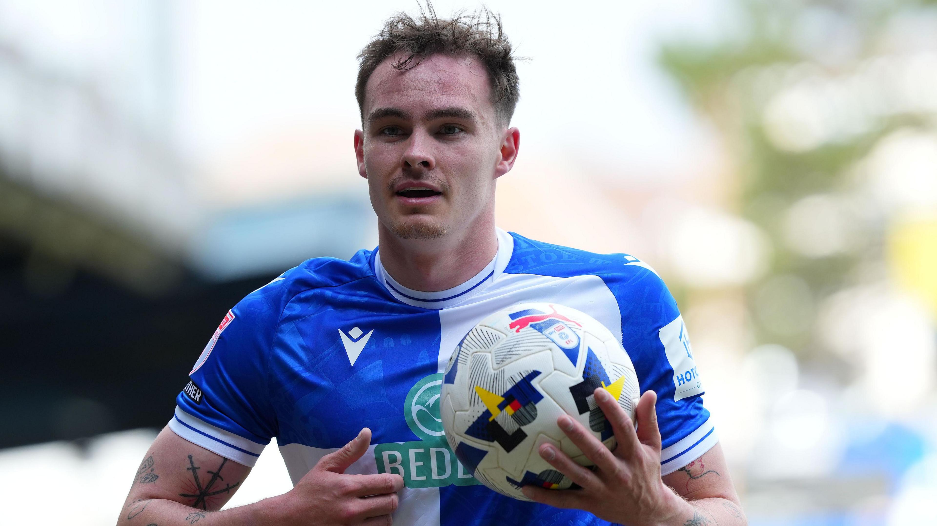 Kofi Balmer holds a football in his right hand while on the pitch during a match for Bristol Rovers this season