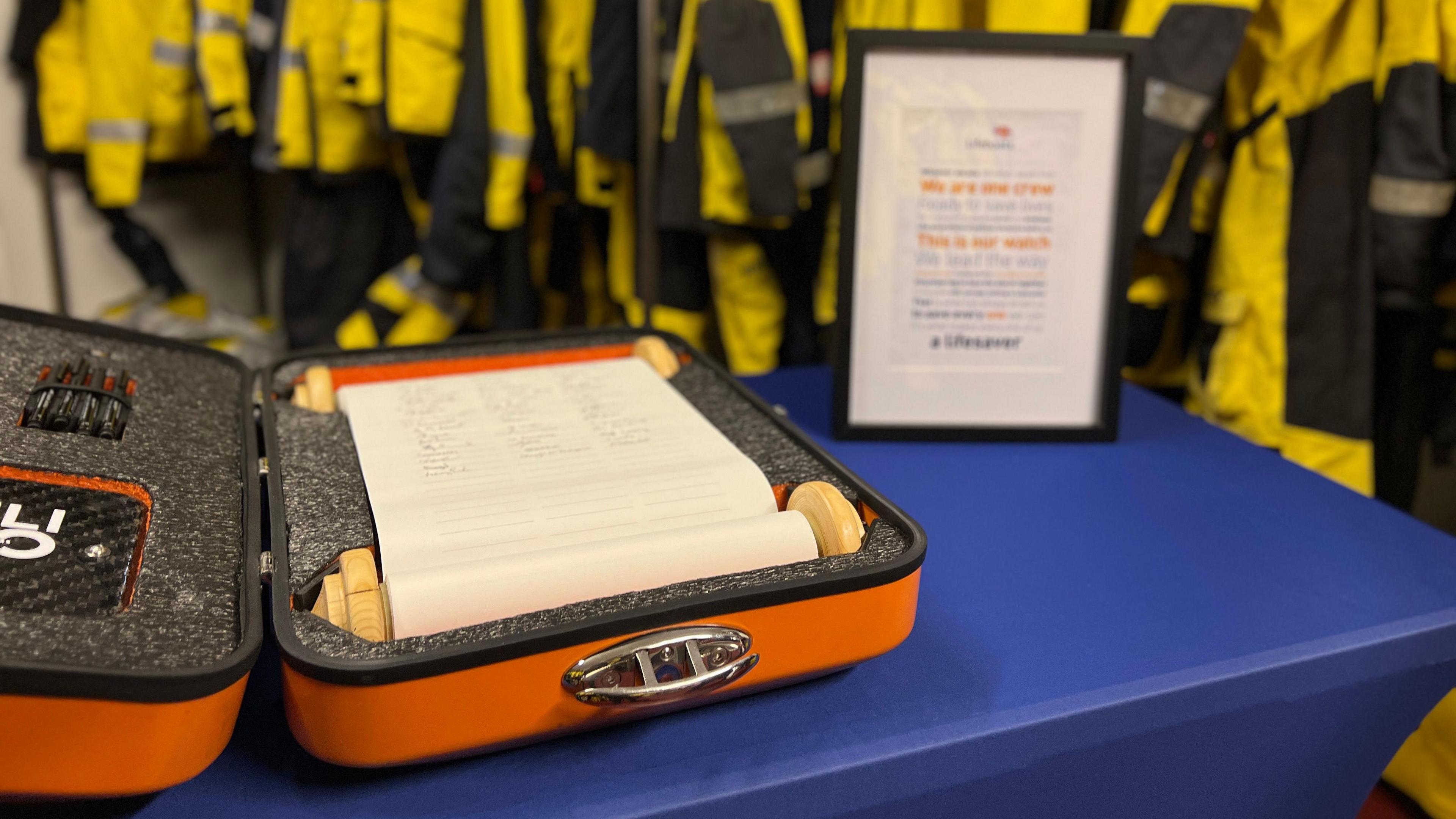 An orange box containing a scroll. The scroll is white and has signatures on it. In the background are a series of yellow waterproofs.