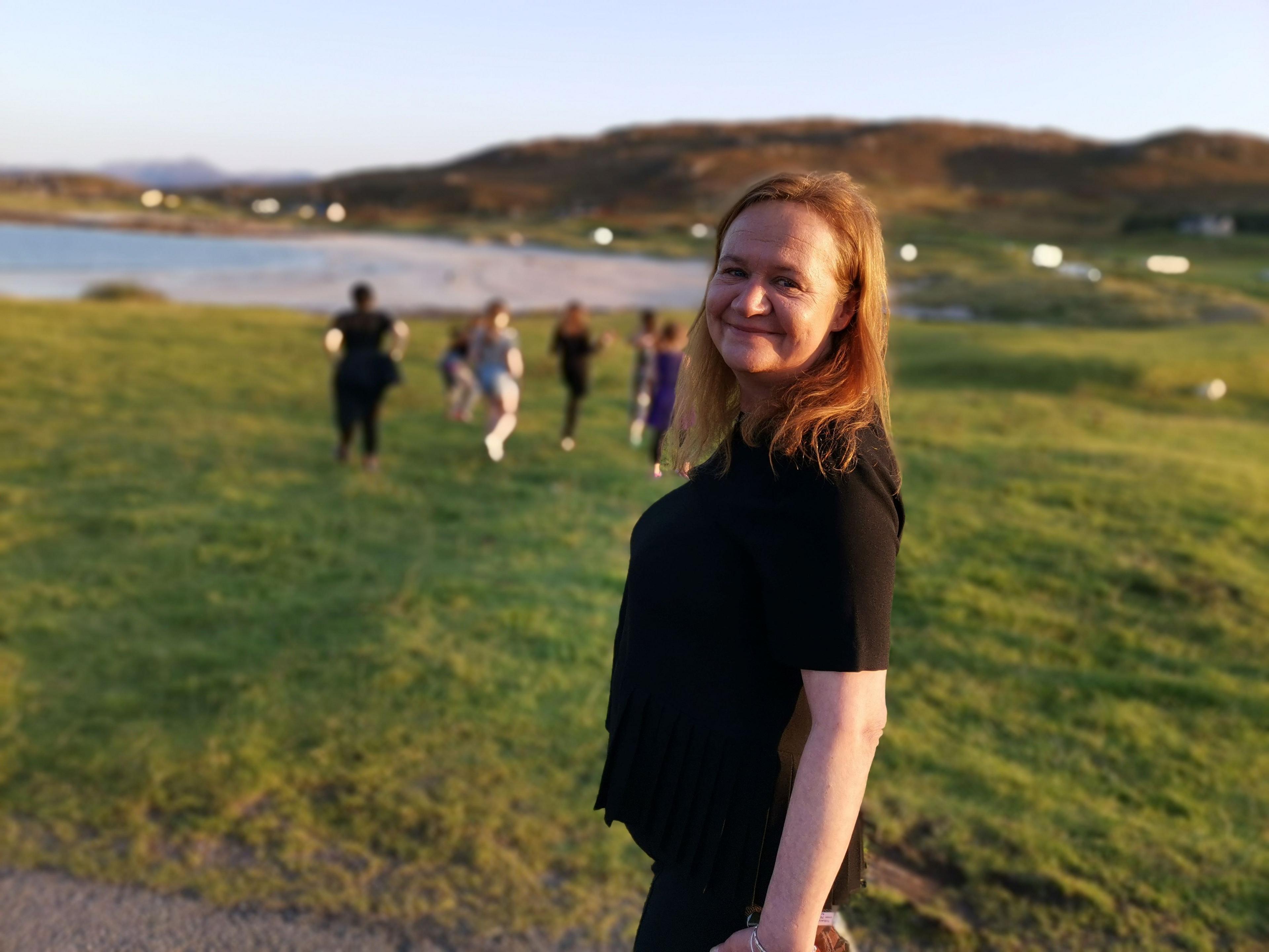 Dr MacRae turning to look at the camera, wearing black clothing. She is standing in front of an area of grass, with Mellon Udrigle beach in the distance. There is a group of young people walking on the grass.