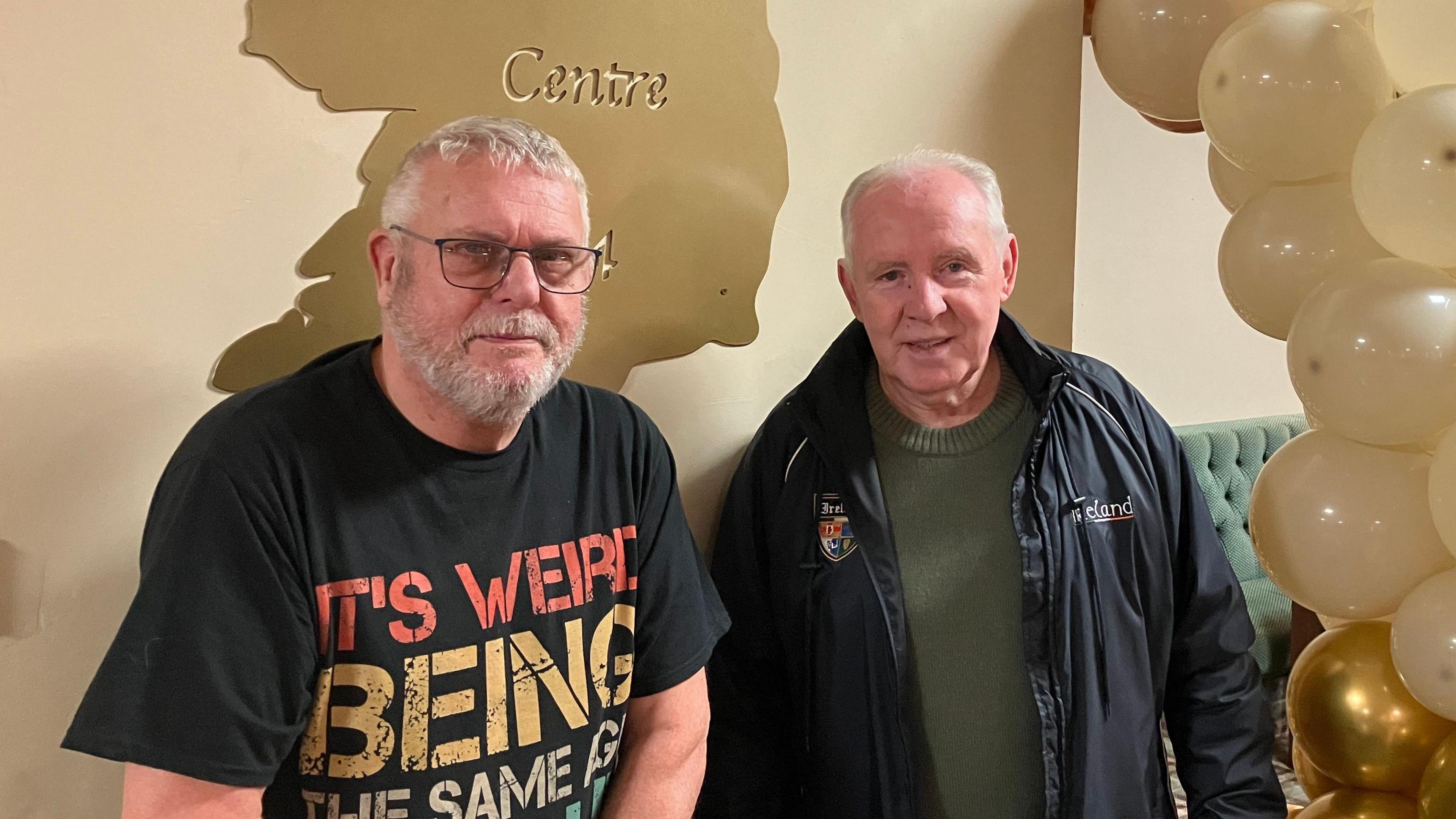 A man wearing a black t shirt and glasses stands next to a man wearing an olive jumper with a black coat. They both have grey hair and are standing inside the Irish Centre. There are lots of white and gold balloons in an arch off to the side, and an metallic gold-coloured map on the wall in the shape of the island of Ireland, with the words "Corby Irish Centre" engraved.