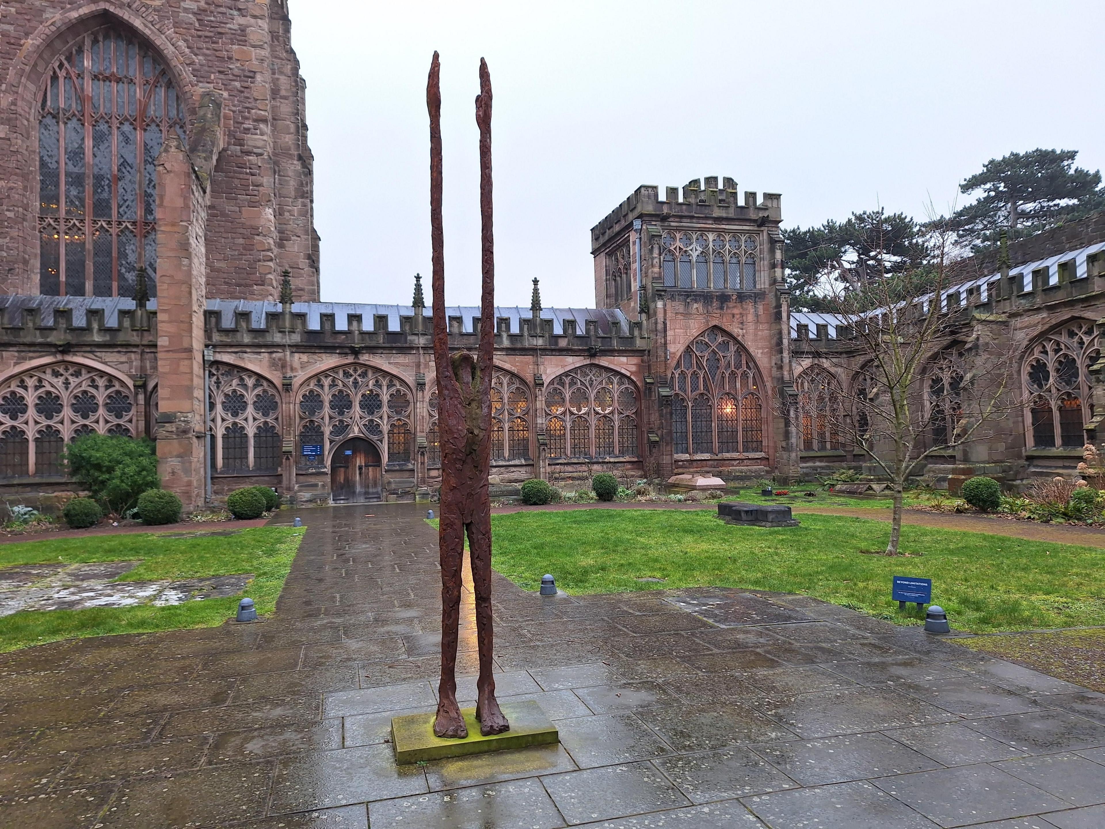 A weathered bronze sculpture of a human figure looking upwards with its hands raised. The very thin, artistic design, has large feet and arms of an exaggerated length, emphasising the sense of reaching to the sky.