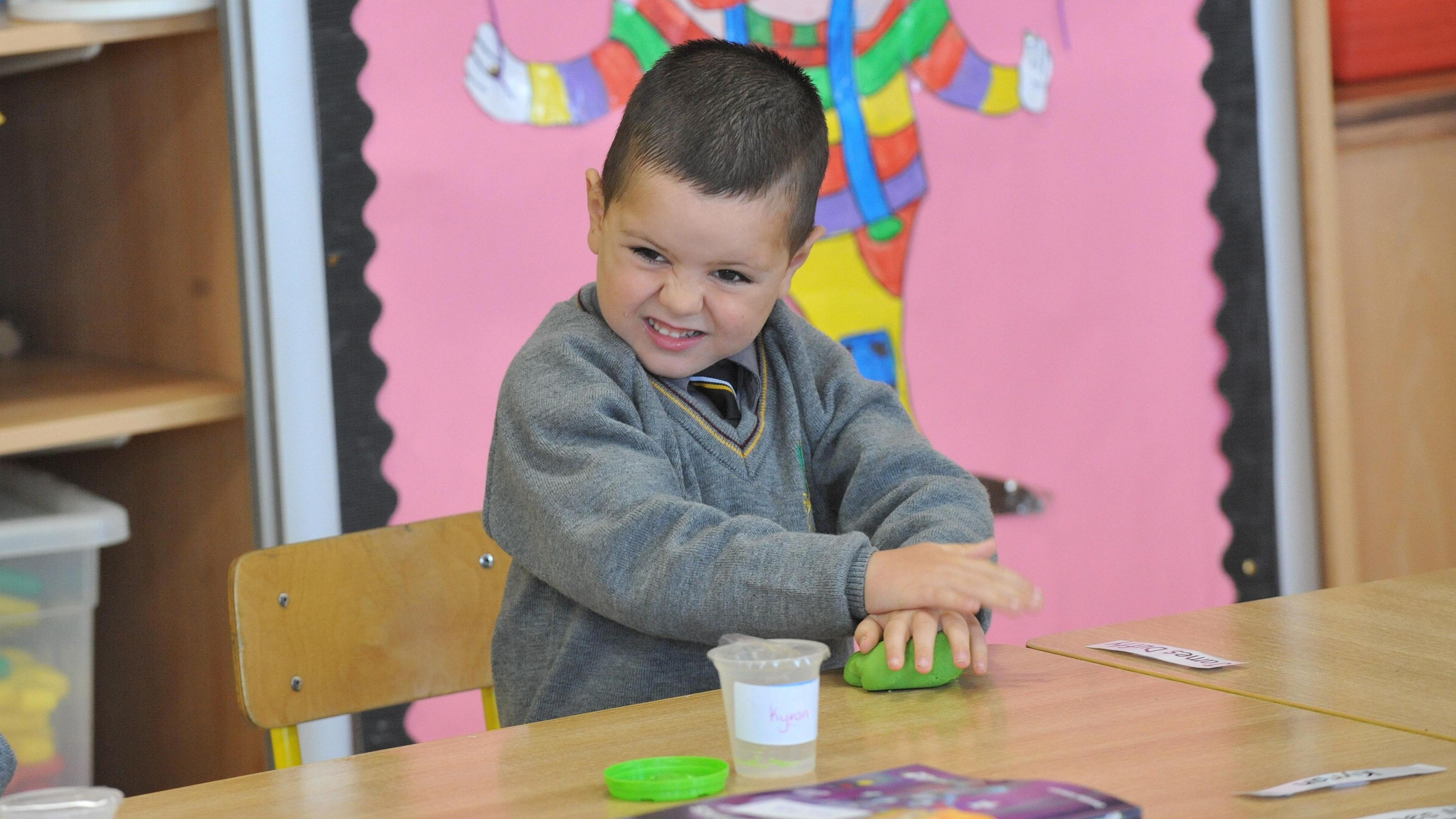 Kyran sits in a school classroom wearing a grey V-neck jumper which has a yellow band around the neck line. He has dark brown hair and brown eyes and his face is scrunched up as he molds some green play dough on the school desk. A small plastic tub bearing his name sits in front of him, with a green lid beside it. Kyran is sitting on a wooden school chair with a large pink classroom display and storage visible behind him. The display shows the lower half of a clowns body, holding a piece of sting which is likely attached to a balloon. 