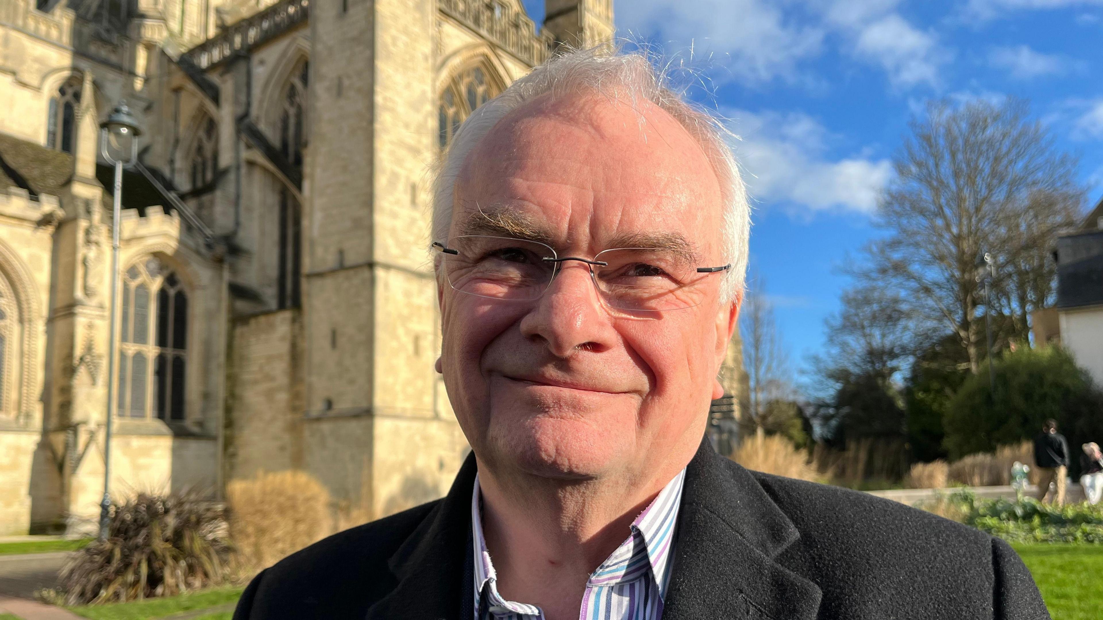 A photo of Gloucester City Council leader Jeremy Hilton standing in front of  Gloucester Cathedral. He has grey hair and is wearing glasses, a striped shirt and a dark jacket