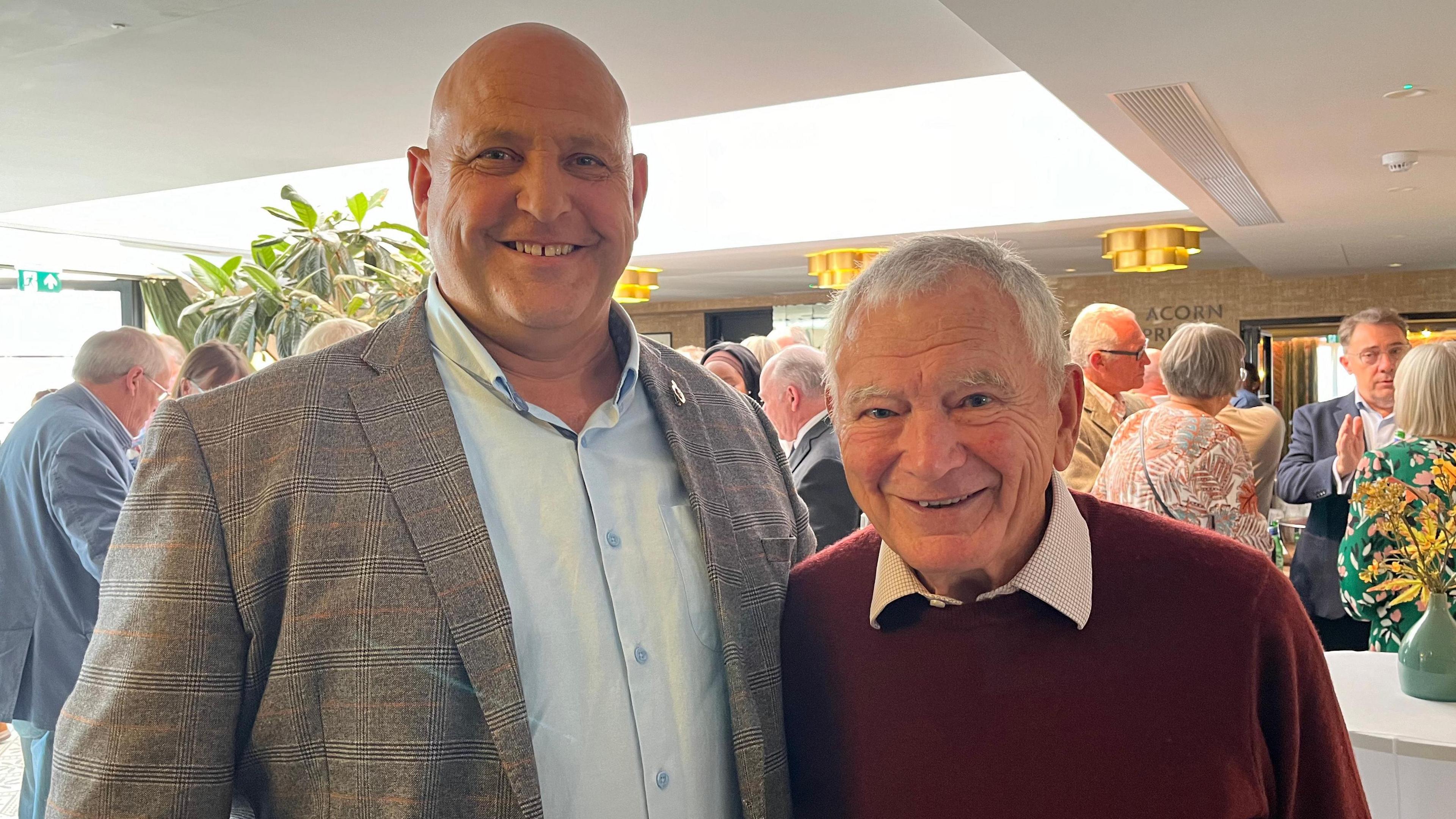 Ben Rowe (left) is a tall bald man in his 40s, wearing a grey blazer and blue shirt. He is smiling. Major Bob Place (right) is a man in his man in his 70s-80s. He has white hair and eyebrows and his smiling showing teeth. He has happy blue eyes.