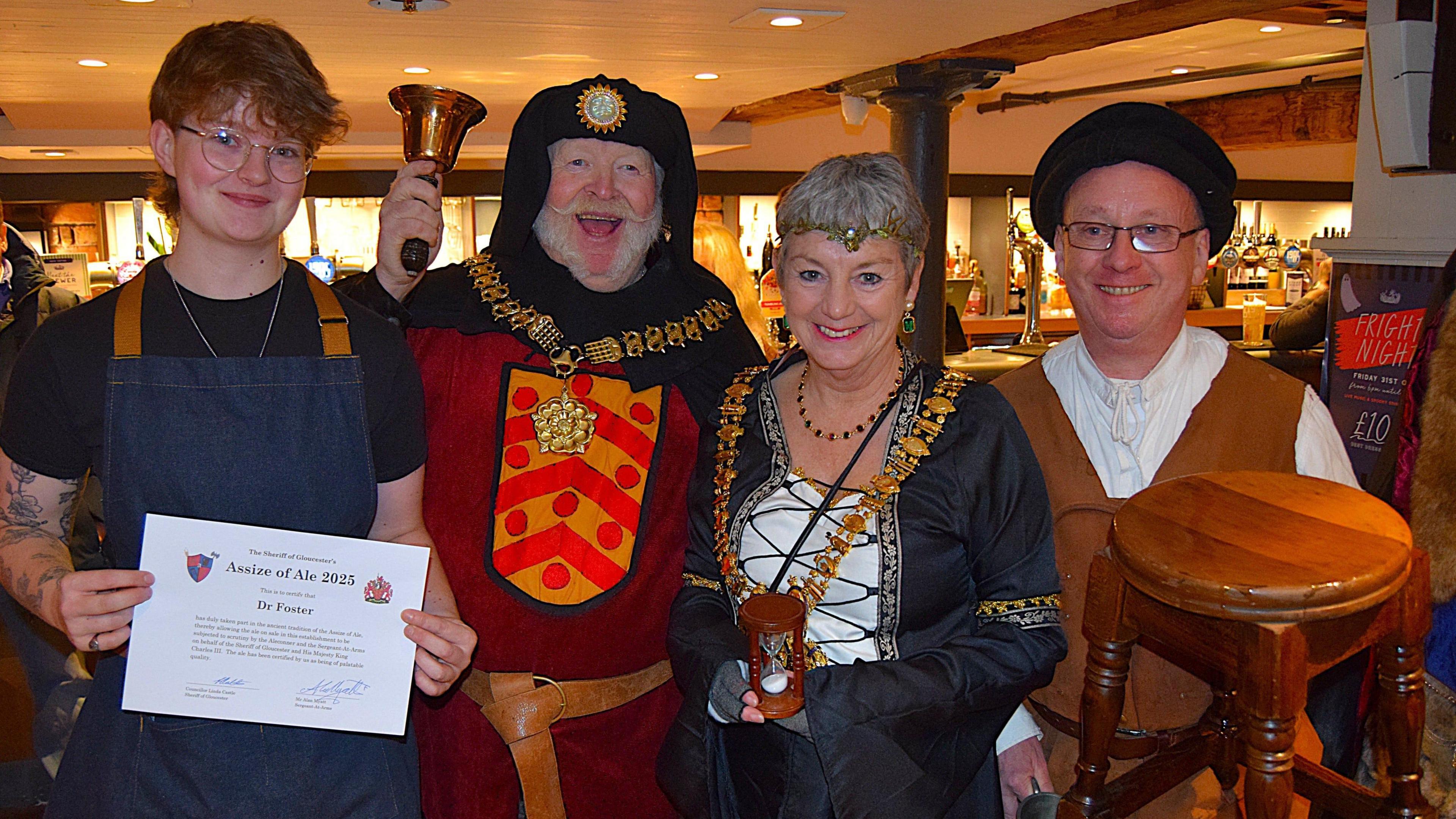 Three people dressed in medieval attire. One is holding a stool, another one a ringing bell. The fourth man on the left is holding a certificate that reads "Assize of Ale".