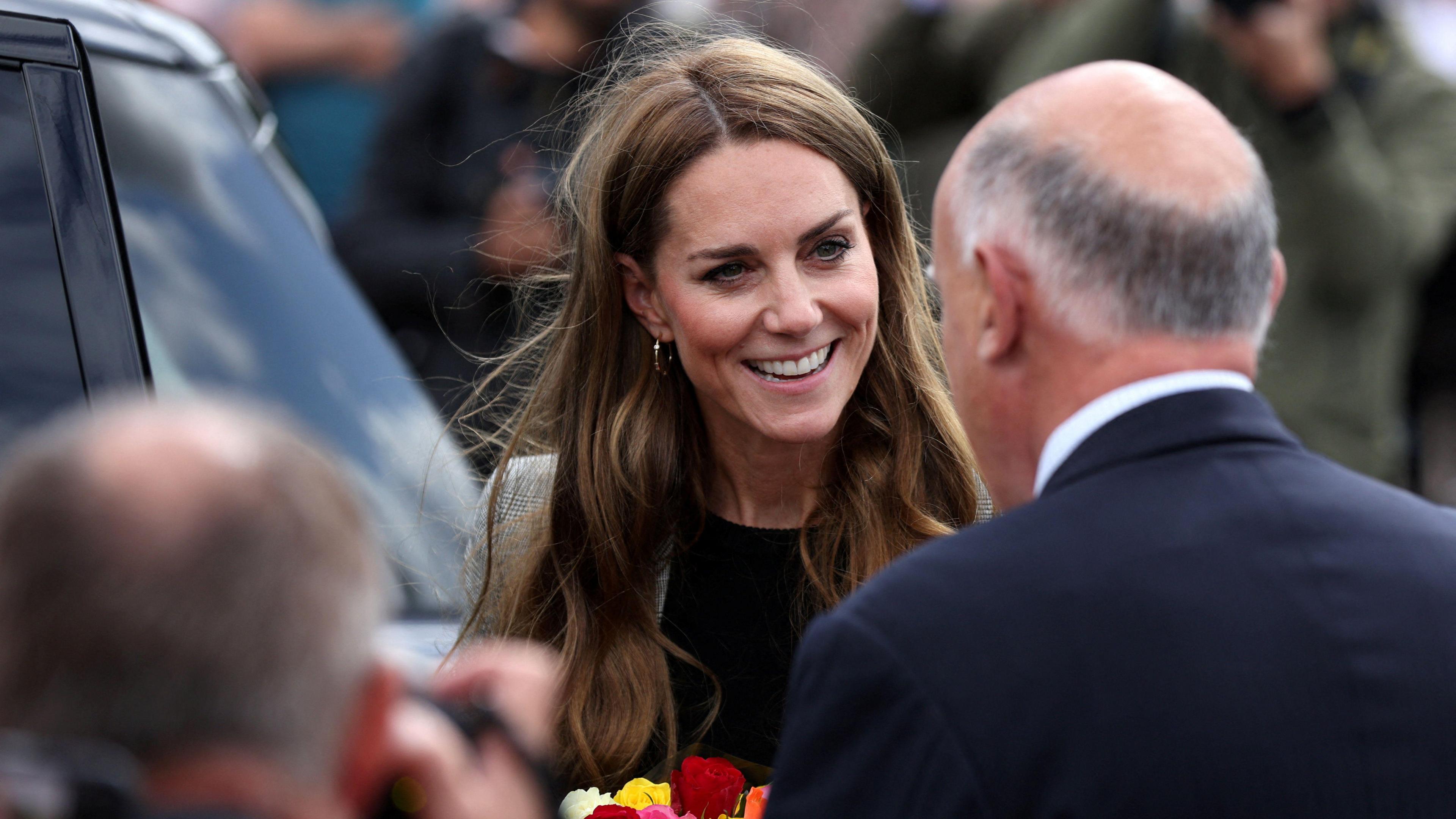 The Princess of Wales smiles and holds brightly coloured flowers whilst speaking to two men with their backs to the camera. She is wearing a black top and brown tweed-style suit and is surrounded by people and standing nearby to a large car.