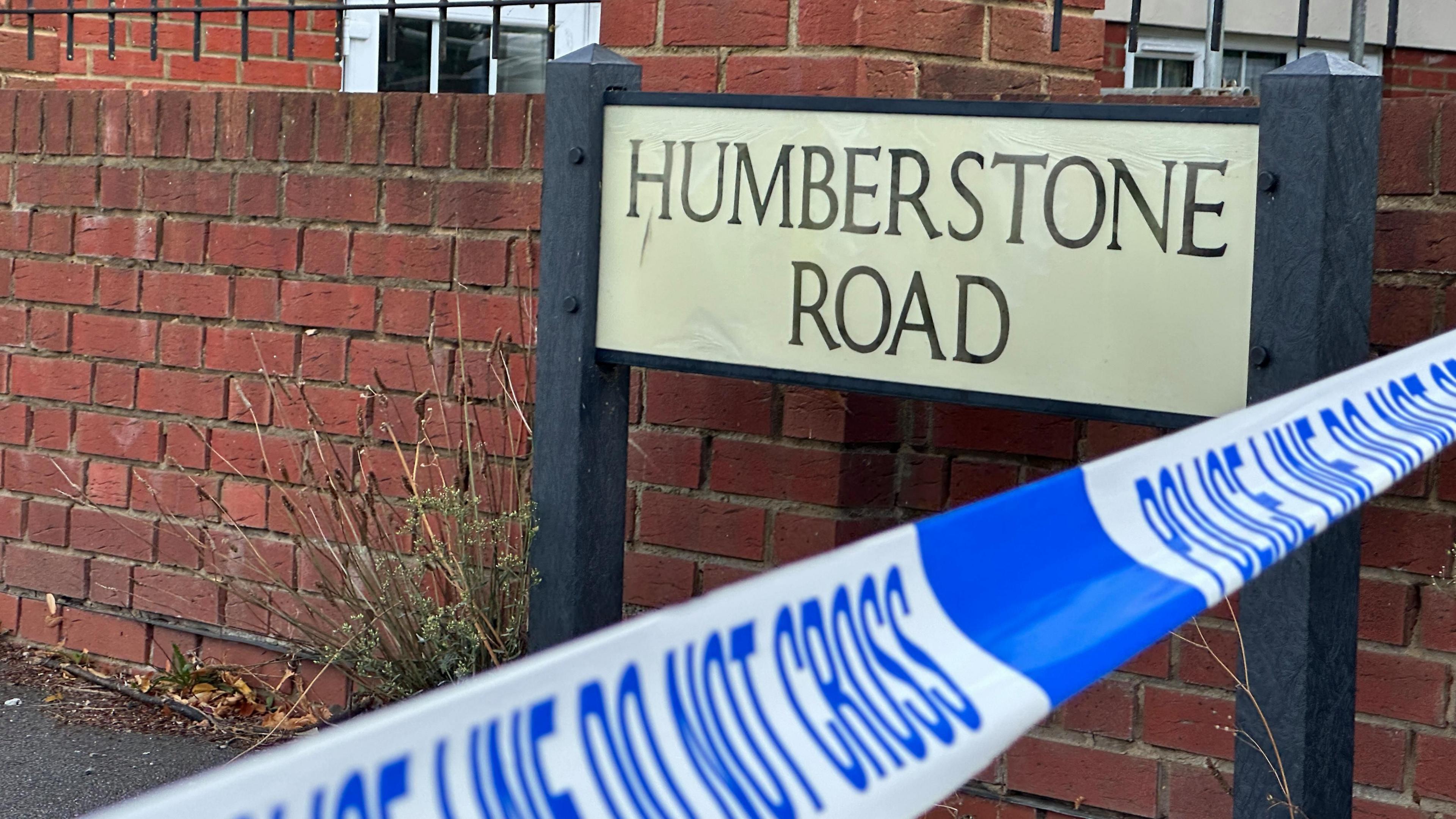 Blue and white police tape which says POLICE LINE DO NOT CROSS is across Humberstone Road in Luton. Within the police tape is a black and white road sign which reads HUMBERSTONE ROAD.