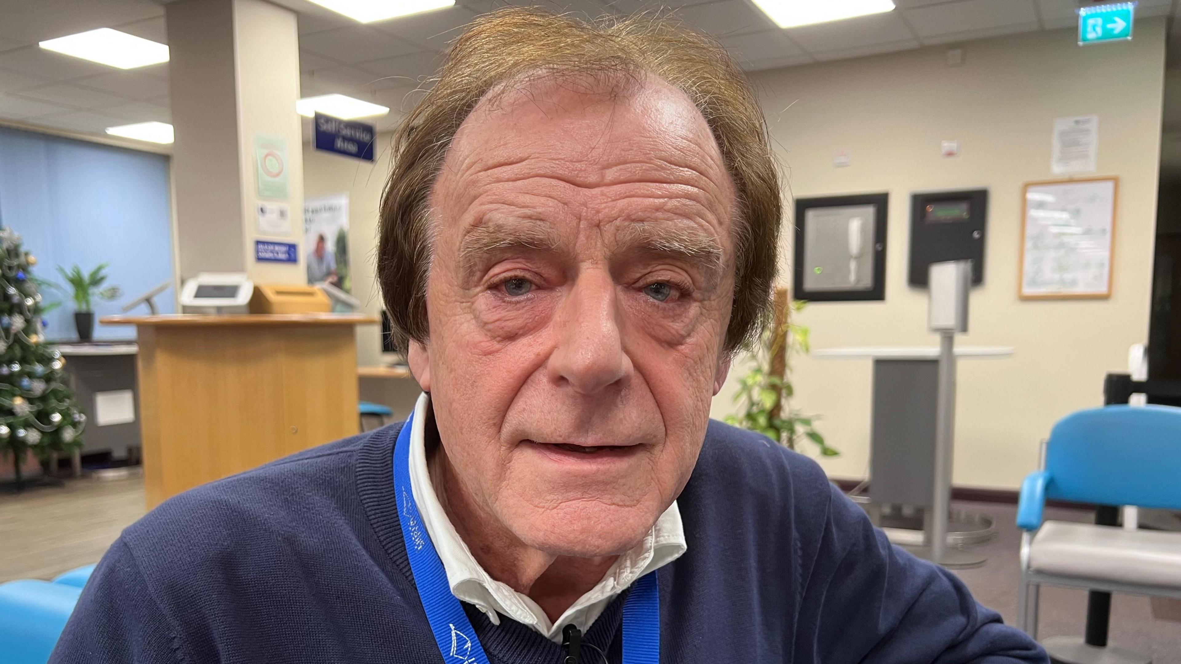 An older man looks at the camera, wearing a white shirt and a blue jumper. He is sat in the entrance of the Isle of Wight council building.