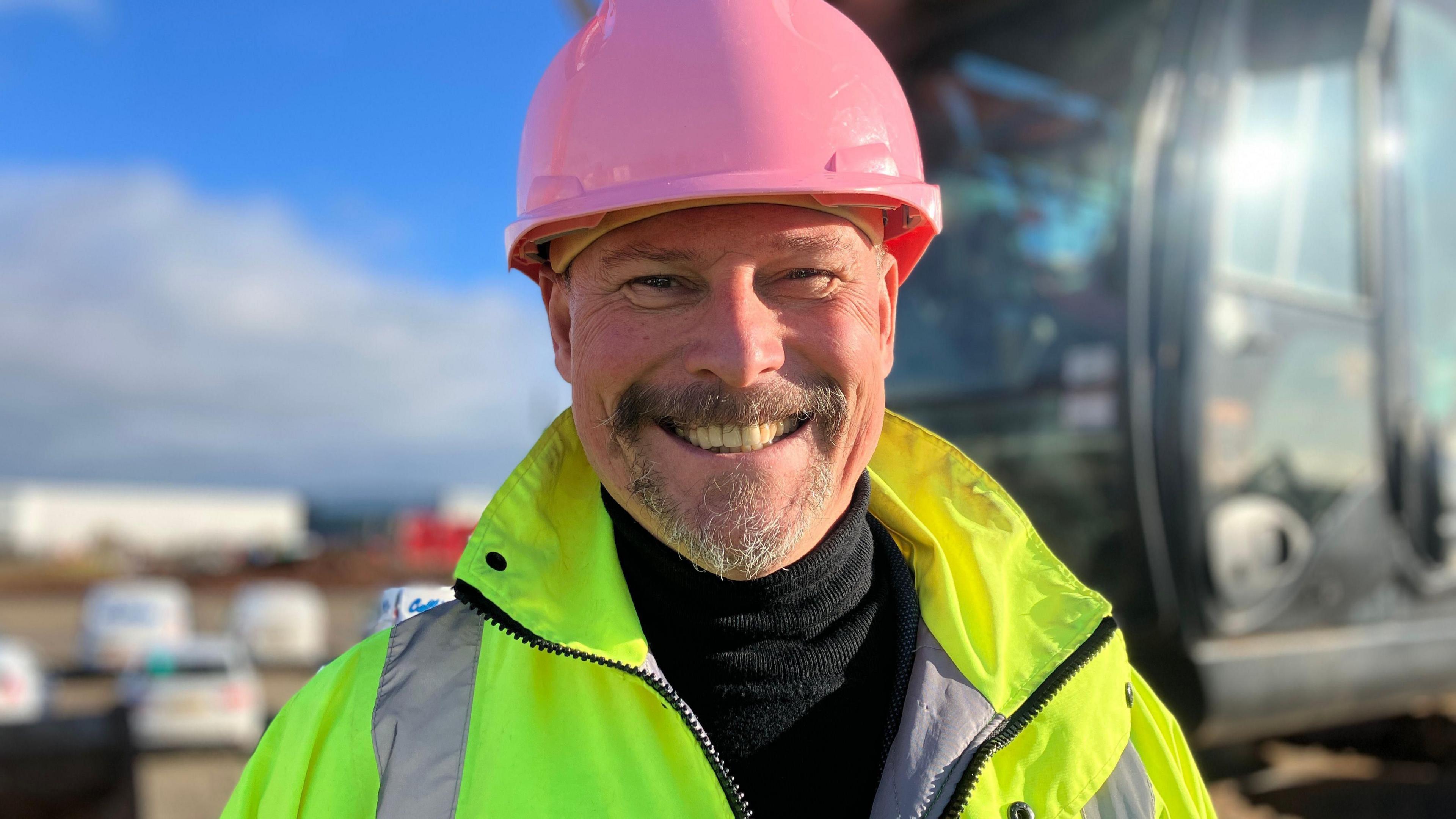 A man with a short grey beard wearing a hi-vis jacket and a pink helmet smiles at the camera.