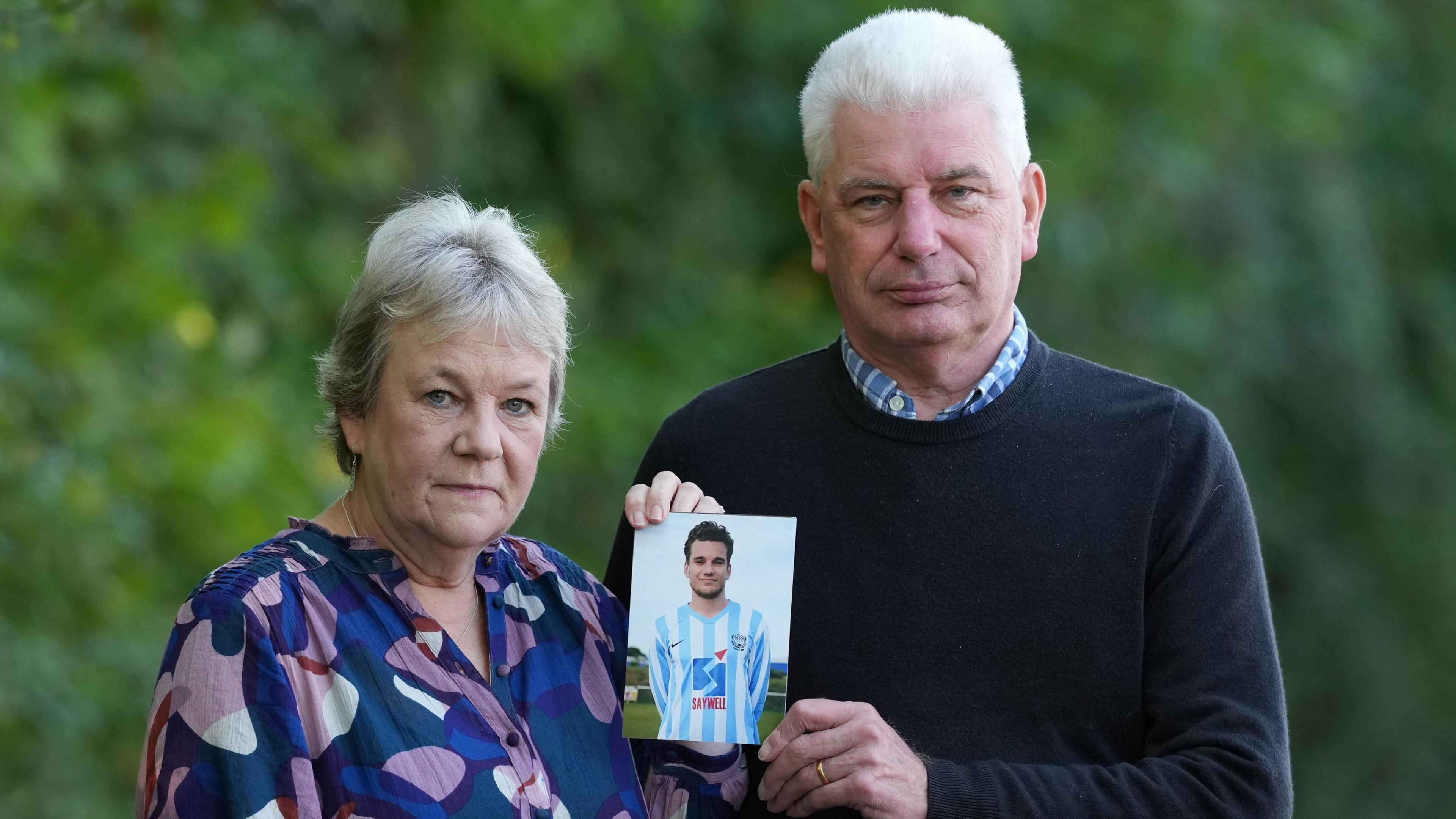 Caroline and Bob Schilt looking at the camera holding a photograph of their son Jacob. They have white hair, Caroline is wearing a multi-coloured blouse and Bob has a striped button-up shirt on under a black jumper