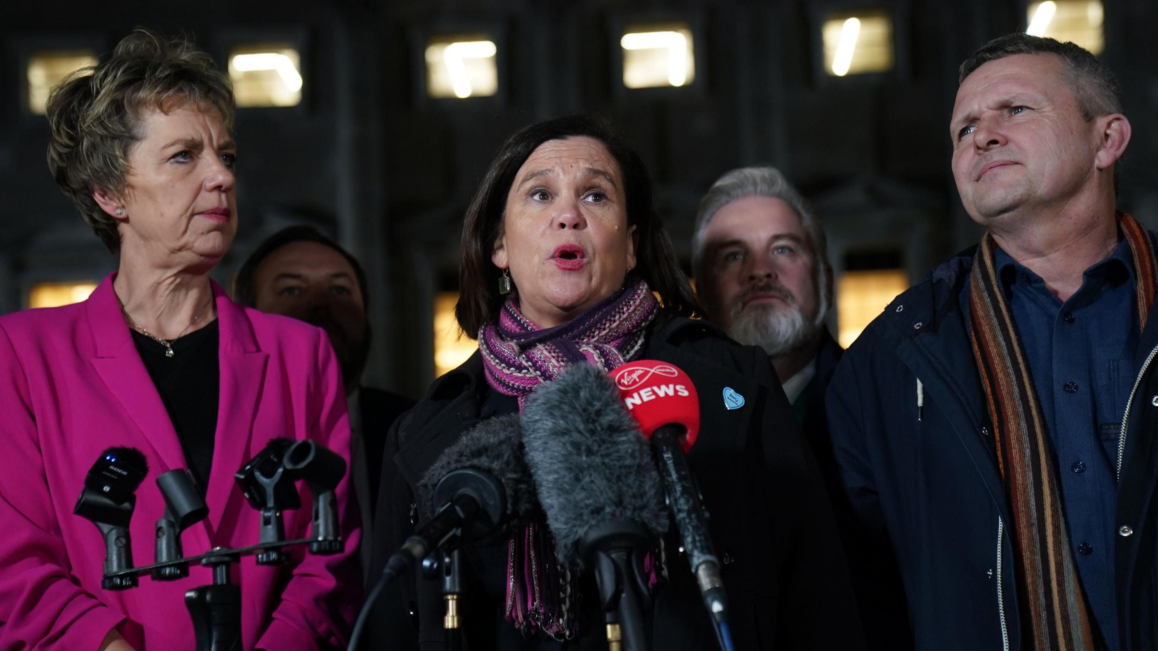 A shot of Labour leader Ivana Bacik in a pink blazer jacket and a black top, Sinn Féin's Mary Lou McDonald in a black coat and patterned scarf and People Before Profit's Richard Boyd-Barrett in a navy coat, navy shirt and striped scarf. They are all standing in a row in front of three microphones in January 2025. 