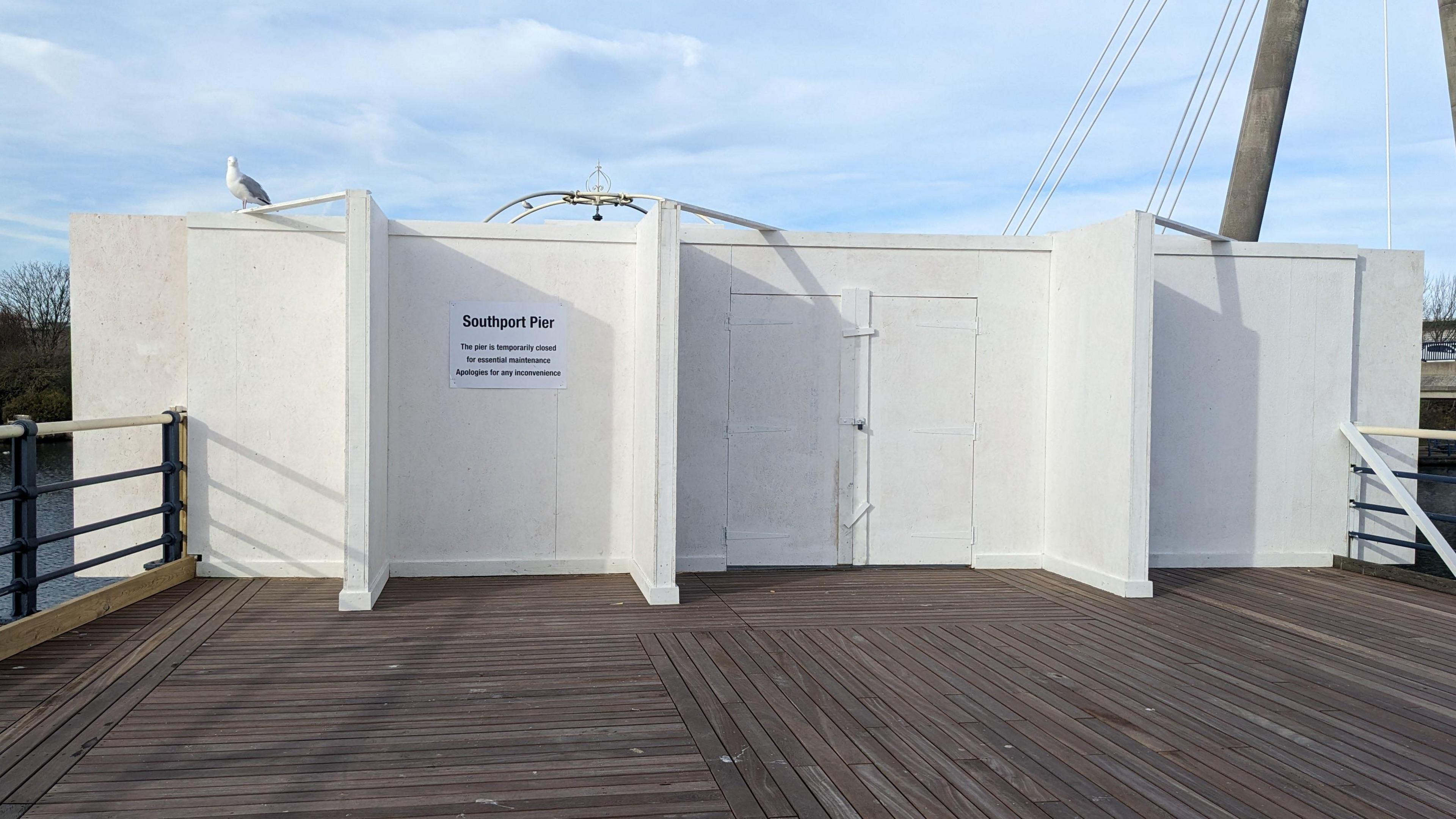 A seagull sits on the white boards across the wooden Southport Pier, with a notice stating it has been shut due to safety issues