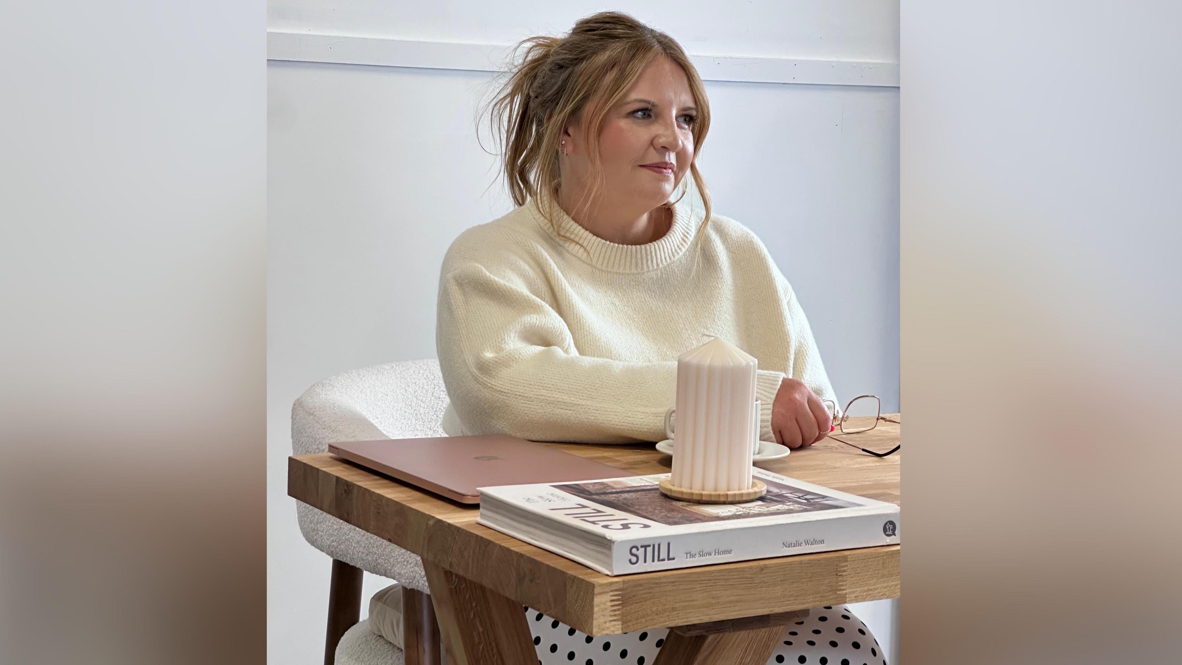 Llun o Sioned yn eistedd wrth ddesg yn edrych bant o'r camera. Mae ganddi wallt melyn a llygaid glas. Mae'n gwisgo siwmper wen. Ar y ddesg o'i blaen hi mae llyfr, cannwyll a gliniadur. Mae'n dal ei sbectol yn ei llaw dde.