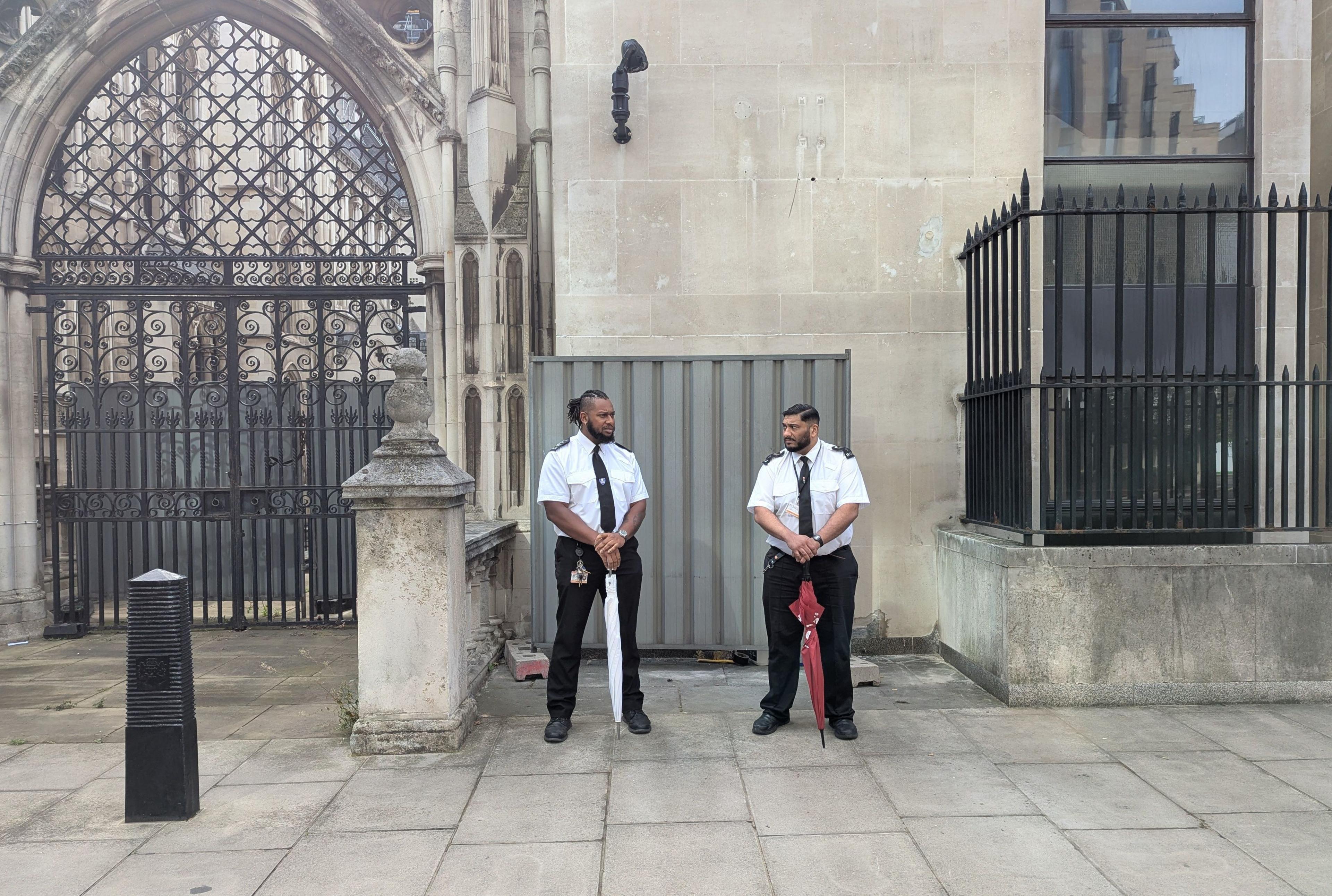 Two men holding furled golf umbrellas in front of a metal barrier in front of a wall. They are dressed in smart security guard outfits and look toward each other.