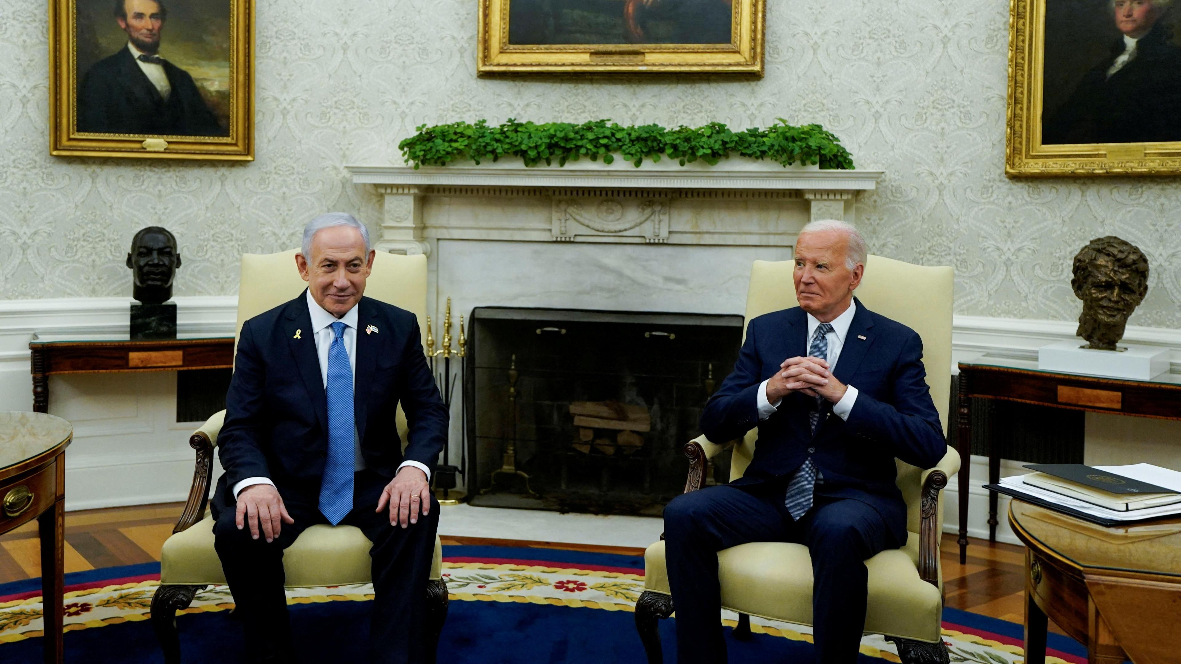 Benjamin Netanyahu sits with his hands on his knees in the oval office, wearing a blue tight and dark suit. Joe Biden sits to his left, wearing a navy suit with clasped hands. 